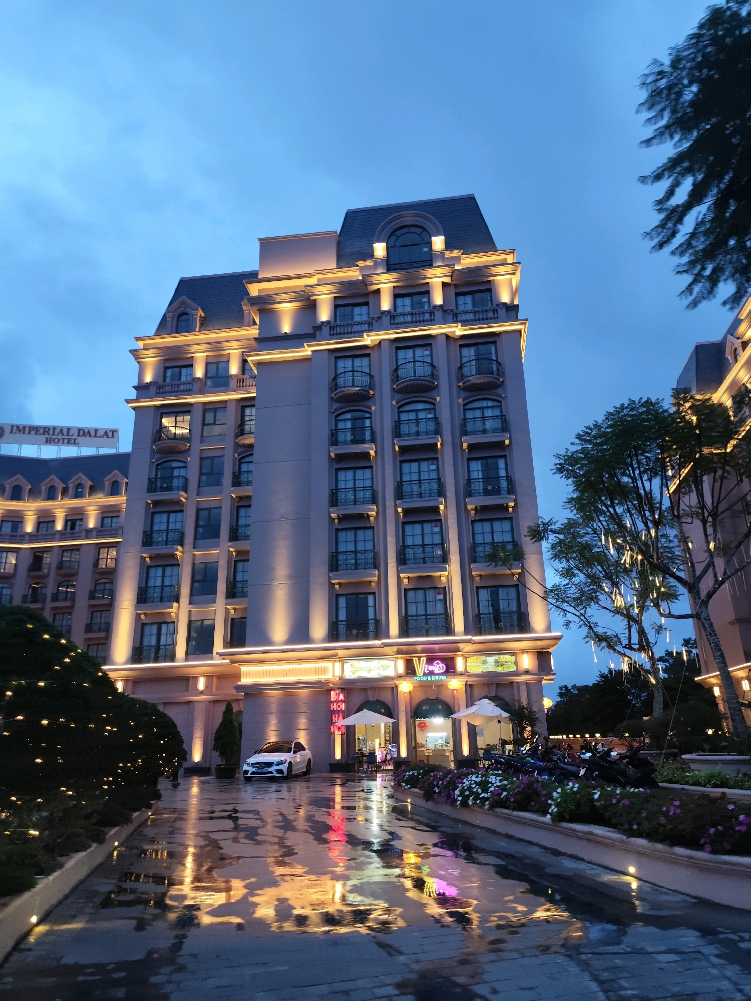 The image depicts the exterior of the Imperial Dalat Hotel at dusk, located in Lâm Viên - Đà Lạt, Vietnam. The hotel building is a tall, multi-story structure with a distinctive mansard roof and illuminated architectural details. A prominent sign reads "IMPERIAL DALAT HOTEL." In front of the hotel, a white car is parked on a wet, reflective surface, suggesting recent rain. The ground is paved with stone and reflects the colorful lights from the building and surrounding trees. A row of flower beds lines a walkway, with various motorcycles parked alongside them. Trees with small, twinkling lights are visible in the background. The sky is a deep twilight blue, with soft clouds. The scene conveys a sense of a well-lit, upscale establishment at the end of the day. There are no visible people in the immediate foreground or mid-ground.