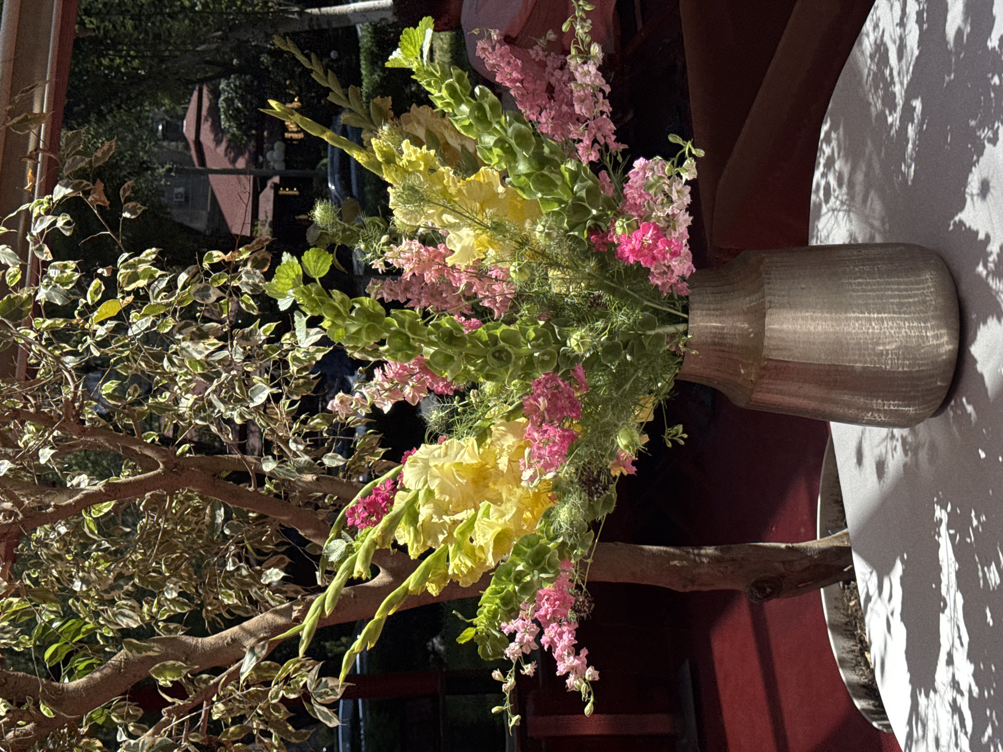 A floral arrangement of pink, yellow, and green flowers is displayed in a metallic vase on a white tablecloth. The vase is positioned on a table in an outdoor setting, with dappled sunlight casting shadows on the tablecloth. Behind the table, a tree with variegated leaves stands in a pot. In the background, a glimpse of buildings and trees suggests an urban environment. The image appears to have been taken outdoors during the daytime. The location is identified as Yerevan, Armenia.
