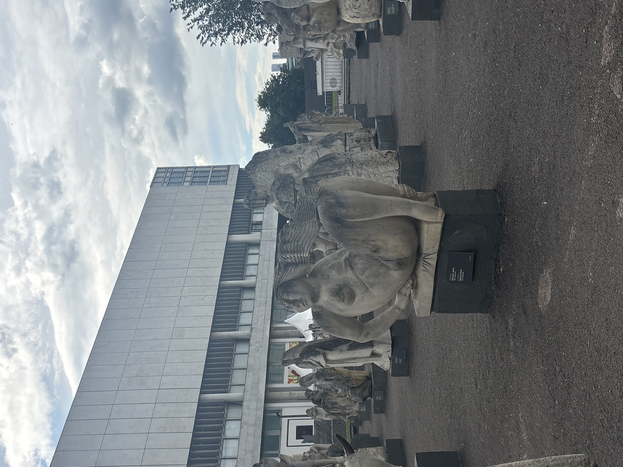 Here's a description of the image:
The image depicts a row of weathered sculptures on pedestals, likely part of an outdoor exhibition. The sculptures appear to be made of stone or a similar material. Behind the sculptures, a tall, modern building with a grid-like facade is visible. The sky is cloudy.
Based on the context clues, particularly the sculptures and architectural style, this image could have been taken in Muzeon Park of Arts, which is located in Moscow, Russia.