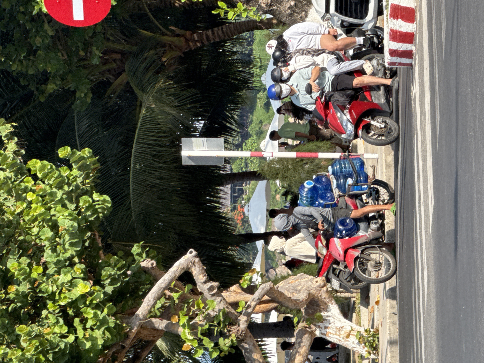 In Bac Nha Trang Ward, Vietnam, on a sunny day, several people are seen on motorbikes near a beach or park area. A prominent red motorbike is stopped at the side of the road, carrying multiple large blue water containers secured to its rear. The driver, wearing a grey long-sleeved shirt, dark shorts, and green flip-flops, has a hood pulled up over his head, obscuring his face. Another motorbike, also red, is positioned in front of the first one. On this second motorbike, four individuals are seated, all wearing masks. The front rider wears a light blue shirt and dark shorts, while the passengers wear a white hoodie, a plaid shirt, and a white hooded jacket respectively. Behind these motorbikes, a white car is visible. The background reveals lush green trees, including palm trees, and several white umbrellas, suggesting a recreational area. A red and white striped barrier is present at the edge of the road. A "no entry" sign is visible on a pole in the background. The ground is paved, with a curb and a pedestrian crossing marked on the road.