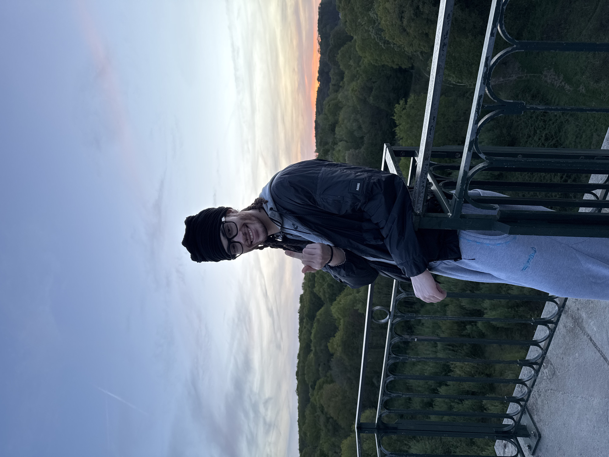 The image captures a young adult standing on what appears to be a high vantage point, possibly a balcony or overlook, above a dense forested valley at sunset. The photograph is oriented vertically, depicting the scene from a tilted perspective.
The subject, likely male, is positioned on the left side of the frame, smiling and looking towards the camera. He wears a black beanie or headwrap, glasses, a black jacket over a grey hoodie, and light grey sweatpants. Dreadlocks are visible peeking from under the beanie. On the left arm of the black jacket, the brand name "RAINS" is clearly visible. On the right leg of the grey sweatpants, there is text written in Arabic script. His right hand is raised, with the thumb and index finger extended in a gesture.
Behind the individual, a panoramic view of a sky painted with sunset hues unfolds. It transitions from soft blues and grays at the top to warm oranges, yellows, and pinks near the horizon, indicating dusk or late evening. Below the colorful sky, a vast expanse of dark green forest covers rolling hills and valleys, stretching into the distance. A dark metal railing with decorative, curved patterns runs across the bottom and right side of the image, separating the viewpoint from the landscape. The ground beneath the railing is a light-colored concrete surface.
The scene conveys a serene and scenic atmosphere, typical of a natural overlook, possibly in Gometz-le-Châtel, France, known for its natural beauty. The person's cheerful expression suggests enjoyment of the view and the moment.