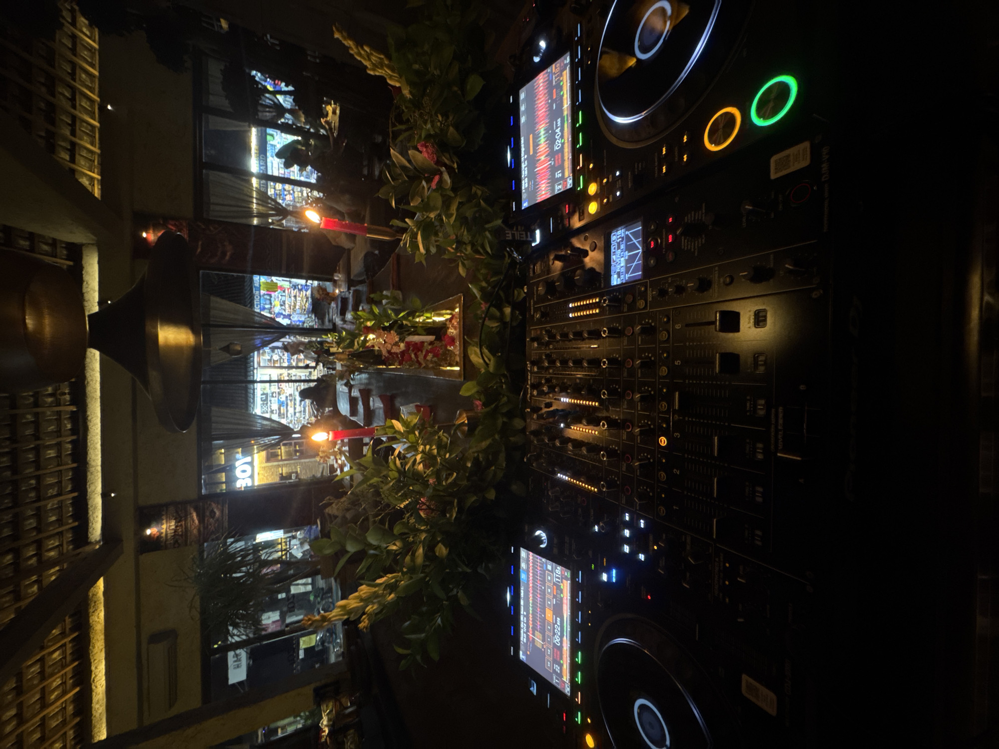 The image captures an indoor scene, possibly within a bar or entertainment venue. Dominating the foreground is professional DJ equipment, specifically a Pioneer DJM-V10 mixer alongside a pair of CDJ media players with lit screens and controls. The equipment is decorated with greenery and flowers, with a lit red candle adding to the ambiance.
In the background, there are blurred figures of people, presumably patrons of the establishment, and a window front exhibiting the store name "BOY". There are also details of the interior, including bamboo-like structures, and ambient lighting. The OCR text suggests that the location could be in North Kuta, Indonesia, based on the appearance of the word "HAR" near the timestamp "06/22", and the number "301" in the background. Additionally, the OCR suggests a location with sections labeled "REVERE" and "TEILE".