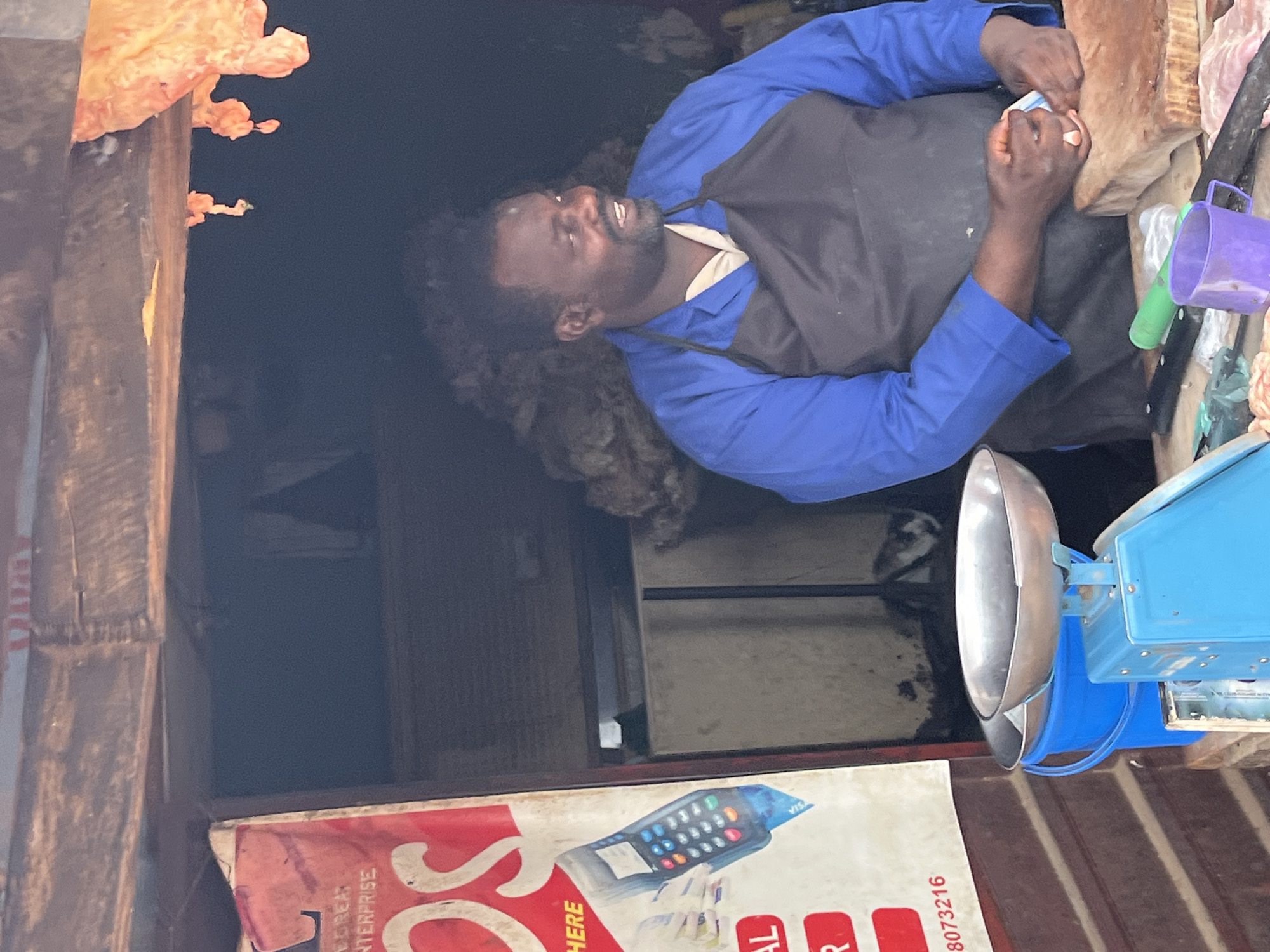 The image depicts a butcher shop in Jos, Nigeria. A man, likely the proprietor, stands behind a counter, wearing a blue long-sleeved shirt and a black apron. He is smiling and looking to his right while holding a piece of meat or a packet in his hands. In front of him is a blue bucket, a silver scale with two pans, and a wooden cutting board with a knife and a purple mug. To the left, a red sign with white lettering is partially visible, advertising an enterprise. The shop appears to be an open-air stall with a wooden roof. The overall scene suggests a busy marketplace environment.