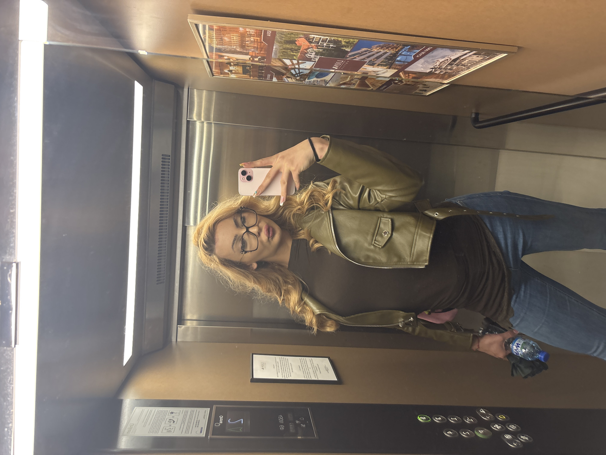 A woman takes a selfie in an elevator in Sofia, Bulgaria. She is wearing glasses, a brown shirt, a green jacket, and blue jeans. She holds a pink cell phone in her left hand and a water bottle and sunglasses in her right. The elevator has metal walls and ceiling, illuminated by fluorescent lights. It also contains an advertisement, a notice, and floor buttons. The floor buttons indicate that the elevator is located at floor 5.