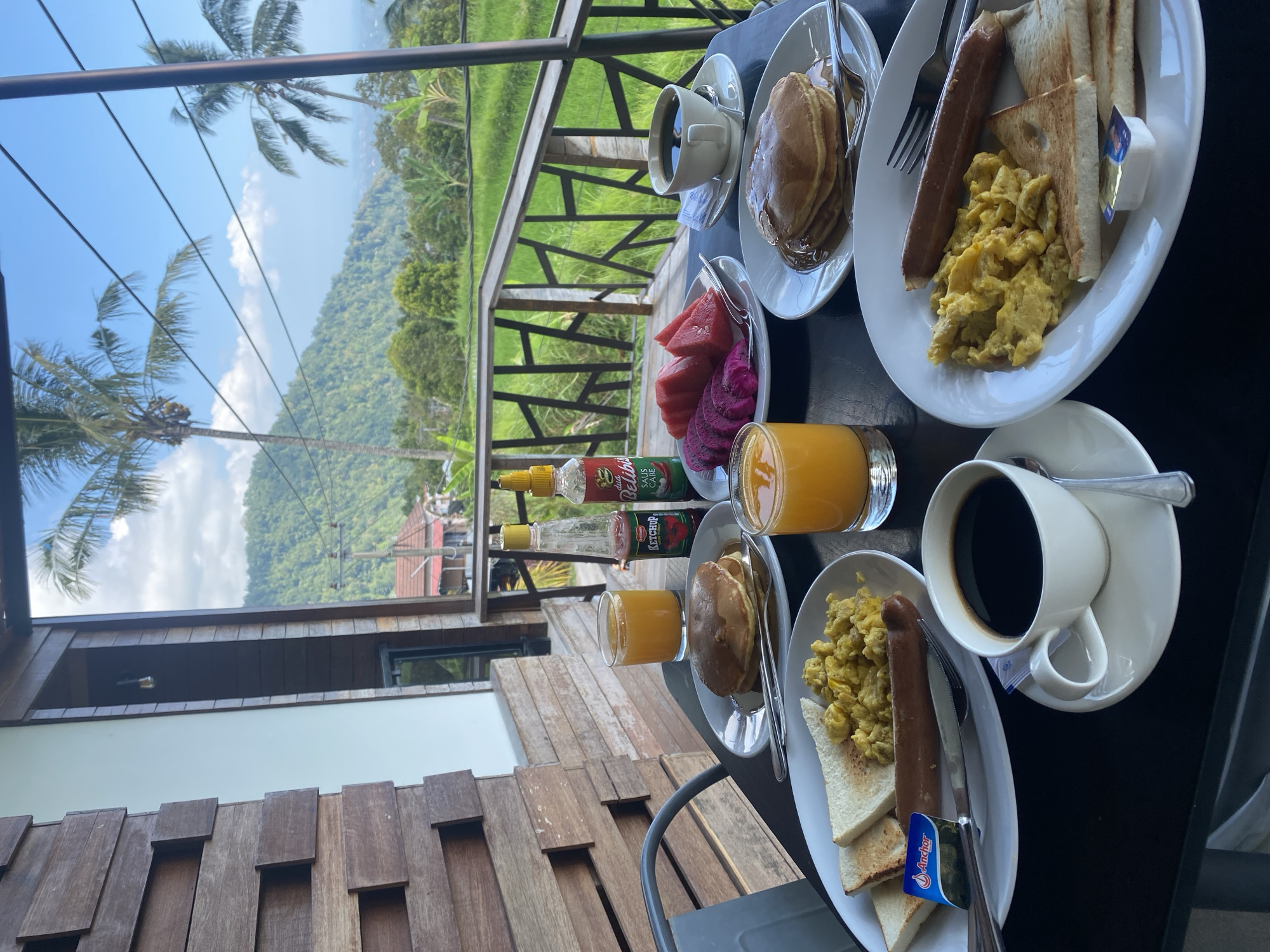 This is a close-up, eye-level, outdoor shot of a breakfast spread on a black table. The table is set on a balcony or patio overlooking a lush green rice paddy field with hills and palm trees in the background. The sky is blue with scattered white clouds. On the table, there are two plates of breakfast. One plate has scrambled eggs, two slices of toast, and a sausage. Next to this plate is a small packet of butter and a cup of black coffee. The second plate has three pancakes with syrup, and a fork and knife. In the background, there are two glasses of orange juice, a bottle of ketchup and a bottle of hot sauce, and a plate of sliced fruit which includes watermelon and dragon fruit. A white building with wooden accents is visible to the left of the frame. The scene appears to be set in Sawan, Indonesia, given the tropical landscape and the visual cues consistent with that region. There are no people visible in the image.