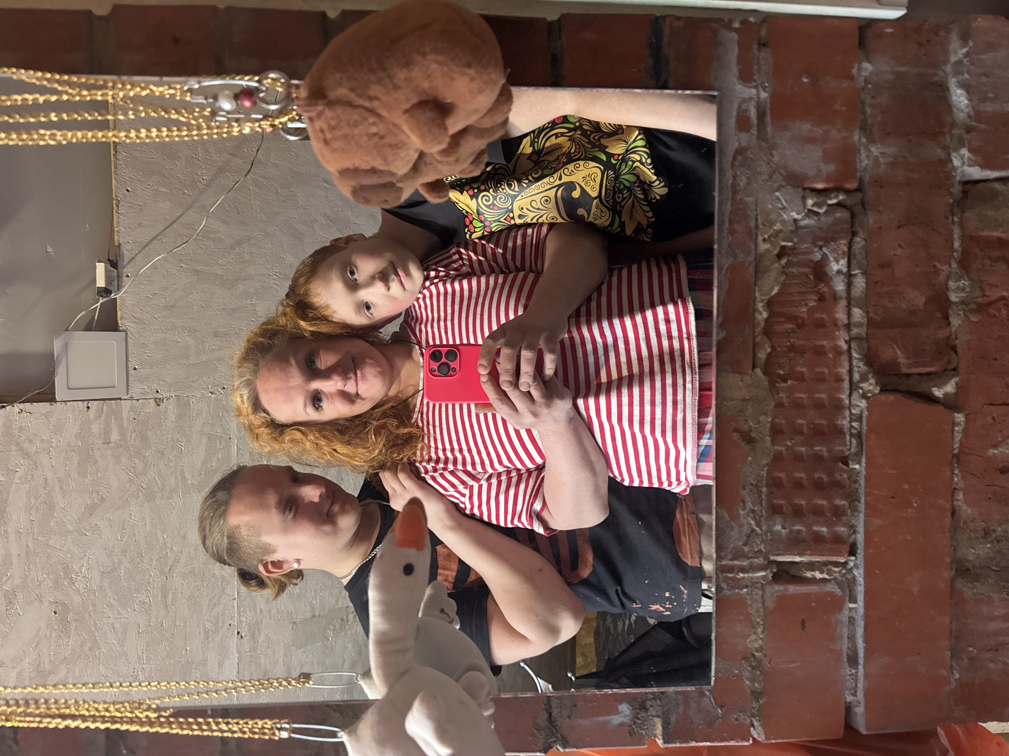 A selfie photograph depicts three individuals and two plush toys, reflected in a mirror. The individuals appear to be related and are posing together. The person at the bottom of the frame has short hair pulled back into a bun, wearing a black T-shirt and a silver chain, with a plush white goose partially obscuring the lower portion of their face. Above them is a woman with curly reddish-blonde hair, wearing a red and white striped shirt, holding a red cellphone as if taking the picture. Above the woman is a child with red hair and a black T-shirt with a yellow floral design, with a brown plush animal above the child's head.
The environment reflected in the mirror includes a grey wall with an electrical outlet and a brick wall that appears to frame the mirror on at least one side. The mirror appears to be hanging from the ceiling, with gold chains visible above the subjects. The location is inferred to be Chaikovsky, Russia based on the user's request.