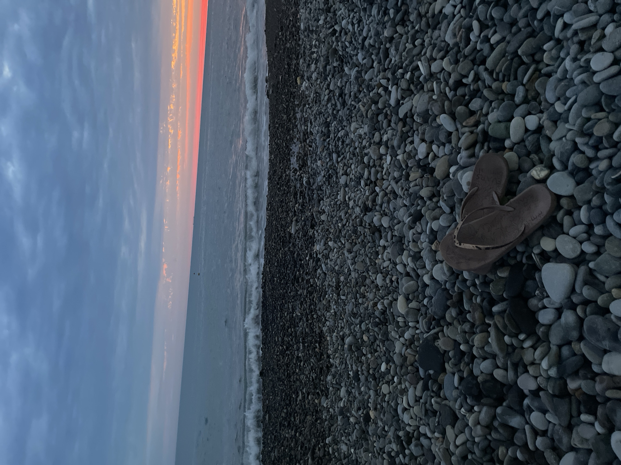 A pair of brown flip-flops rests on a dark, pebble-covered beach. The pebbles are of various sizes and shades of gray and black. In the background, the ocean meets the shore with gentle waves. The sky above is a dramatic mix of dark, cloudy blue with a sliver of vibrant pink and orange from a sunset visible on the horizon. The overall atmosphere is serene and melancholic.