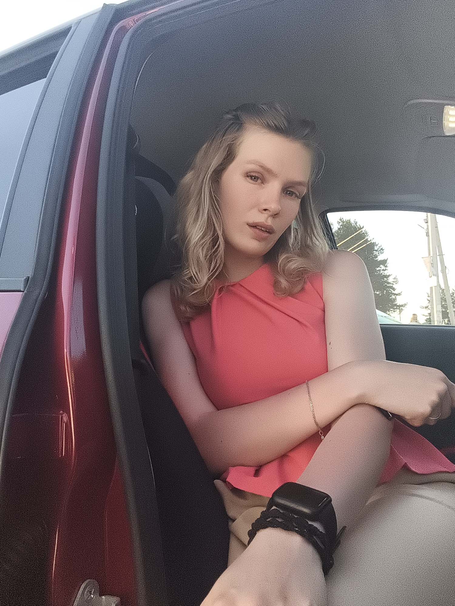 A person with fair skin and wavy blonde hair sits inside a red car. They are wearing a sleeveless pink top and light-colored pants. A black smartwatch is visible on their left wrist, along with a delicate gold bracelet on the other arm. The car door is open, and a glimpse of the outdoors suggests it could be a residential area, possibly in Ягул, Russia, although there is no definitive evidence to confirm the specific location.