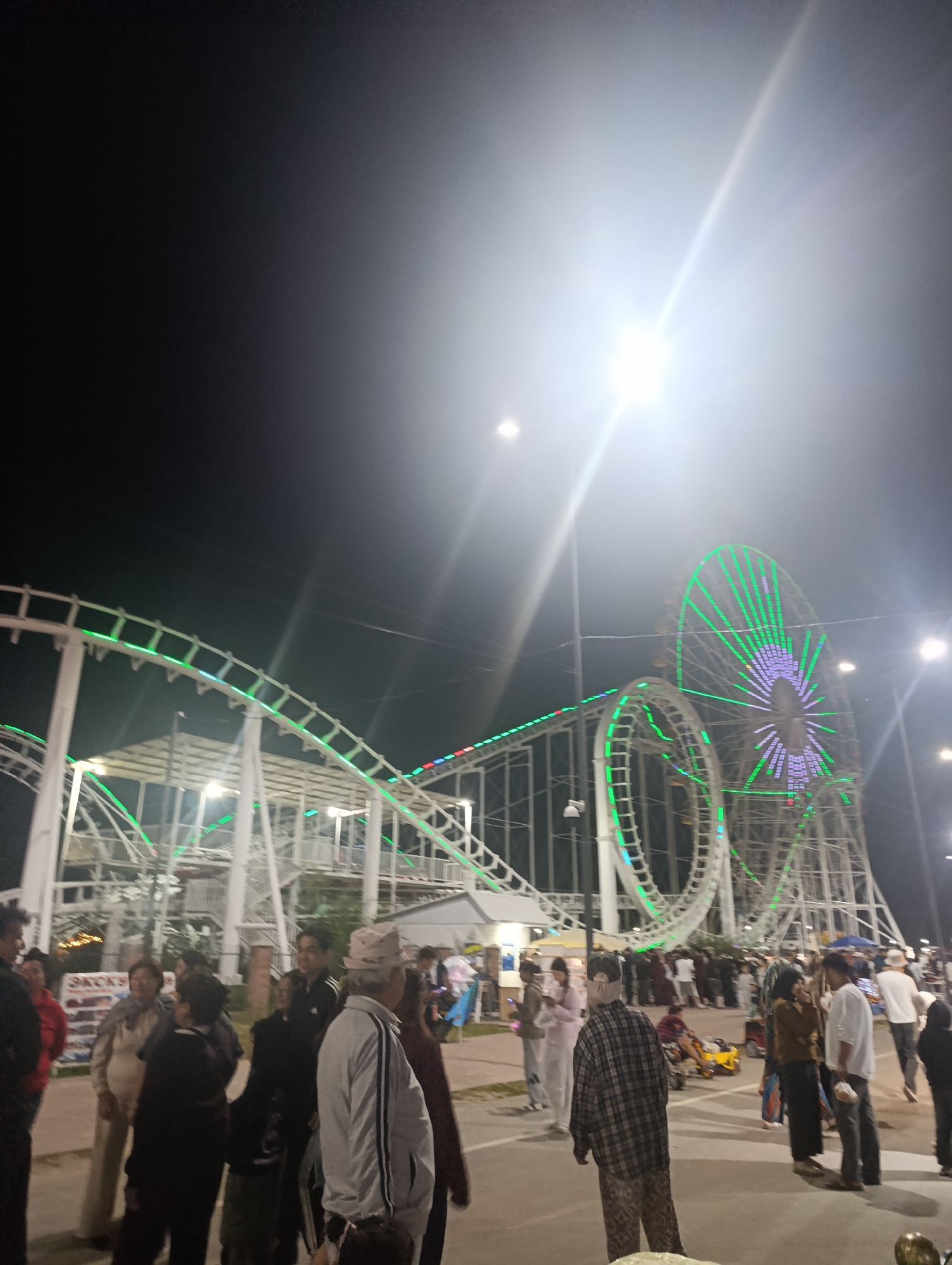 The image depicts an amusement park at night, likely in the city of Bosteri, Kyrgyzstan. A white roller coaster with green and red lights winds through the scene, with a large Ferris wheel illuminated in green and purple to its right. Several people are visible, some standing in groups and others walking. The ground appears to be paved, and there are streetlights and other park infrastructure present. The overall atmosphere is that of a lively evening at an amusement park.