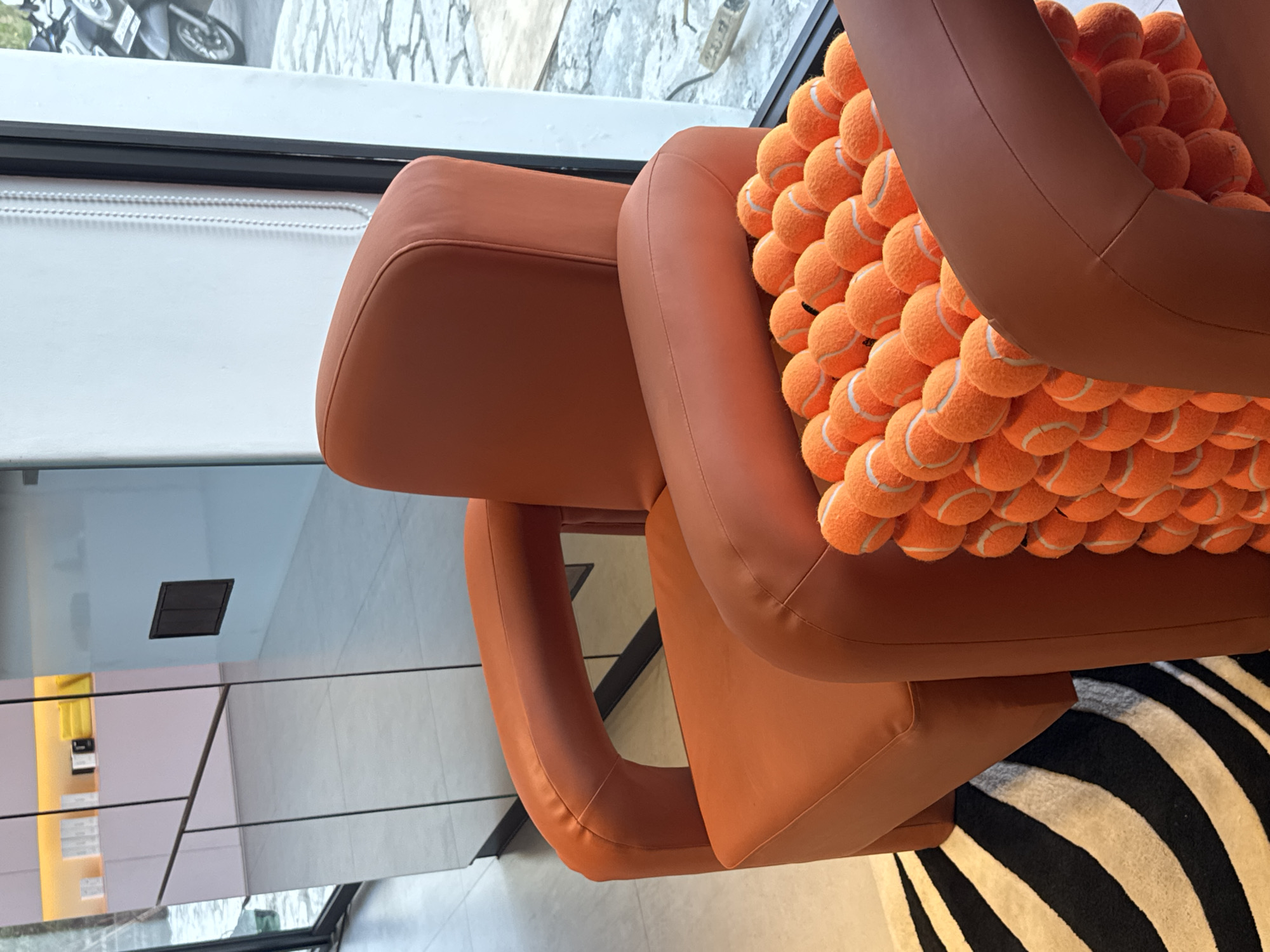 The image shows an interior scene with modern furnishings. In the foreground, a plush orange armchair is prominently featured, with its armrest curving upwards. The chair is partially filled with a stack of bright orange tennis balls, creating a textured and visually interesting element. To the left of the armchair, a portion of a black and white striped rug is visible, adding a bold pattern to the floor.
Behind the armchair, a large reflective surface, likely a mirrored cabinet or wall, dominates the background. This surface reflects the surrounding environment, including what appears to be a display of minimalist white boxes and yellow objects on shelves above. A dark rectangular vent or niche is also visible on a wall reflected in the mirror.
To the right of the armchair, a large window allows natural light to enter the space. Through the window, a glimpse of an outdoor area can be seen, featuring a paved surface with an irregular pattern and what appears to be a parked scooter or motorcycle. The lighting suggests it is daytime. The overall aesthetic is modern and stylish, possibly a retail space, a lounge area, or a contemporary home. The scene does not depict any individuals.