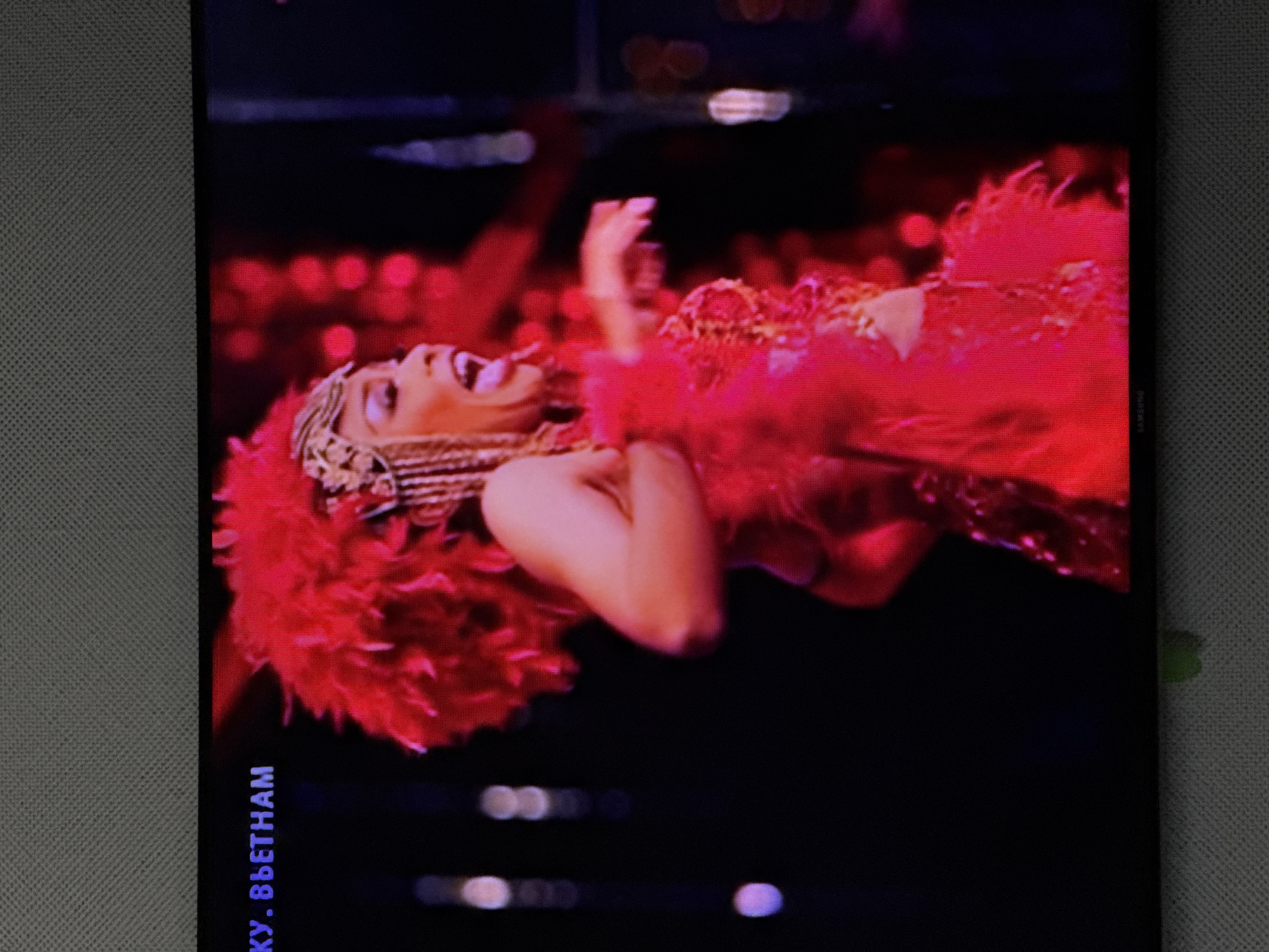 A woman in a red feathered costume and headdress is performing on a stage. The background is dark with blurred red lights. The woman is looking to her right with her mouth open. She is wearing a necklace and a sparkling bodice. The image is a photograph of a television screen. Text is visible in the top left corner of the screen, which reads "КУ. ВЬЕТНАМ". The bottom of the screen displays the Samsung logo. The location is not specified in the image.
