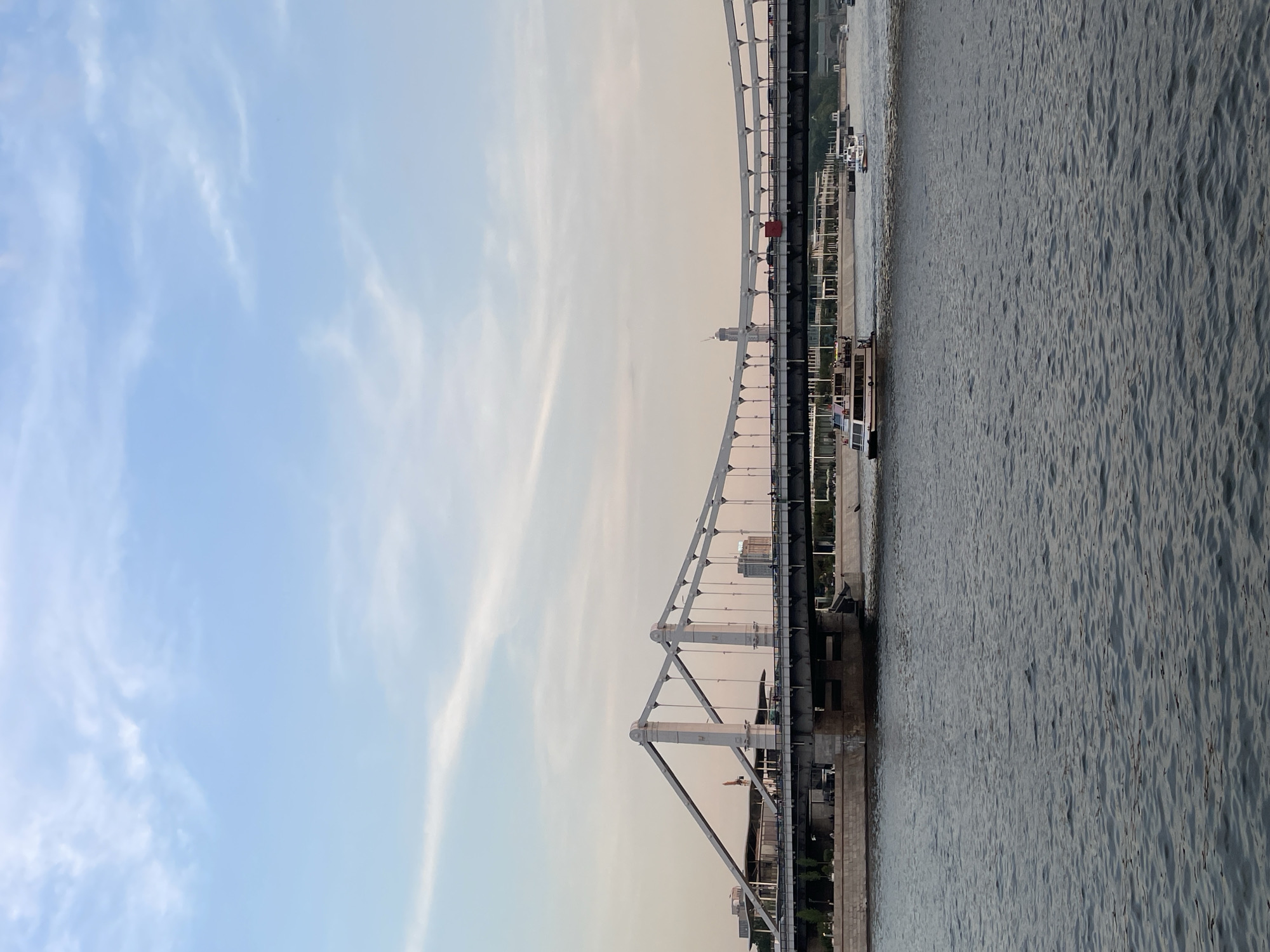 The image shows the Crimean Bridge in Moscow, Russia. It is a steel suspension bridge that spans the Moskva River. The sky is visible in the upper left of the image, and the river is visible on the right. Several boats are visible on the river.