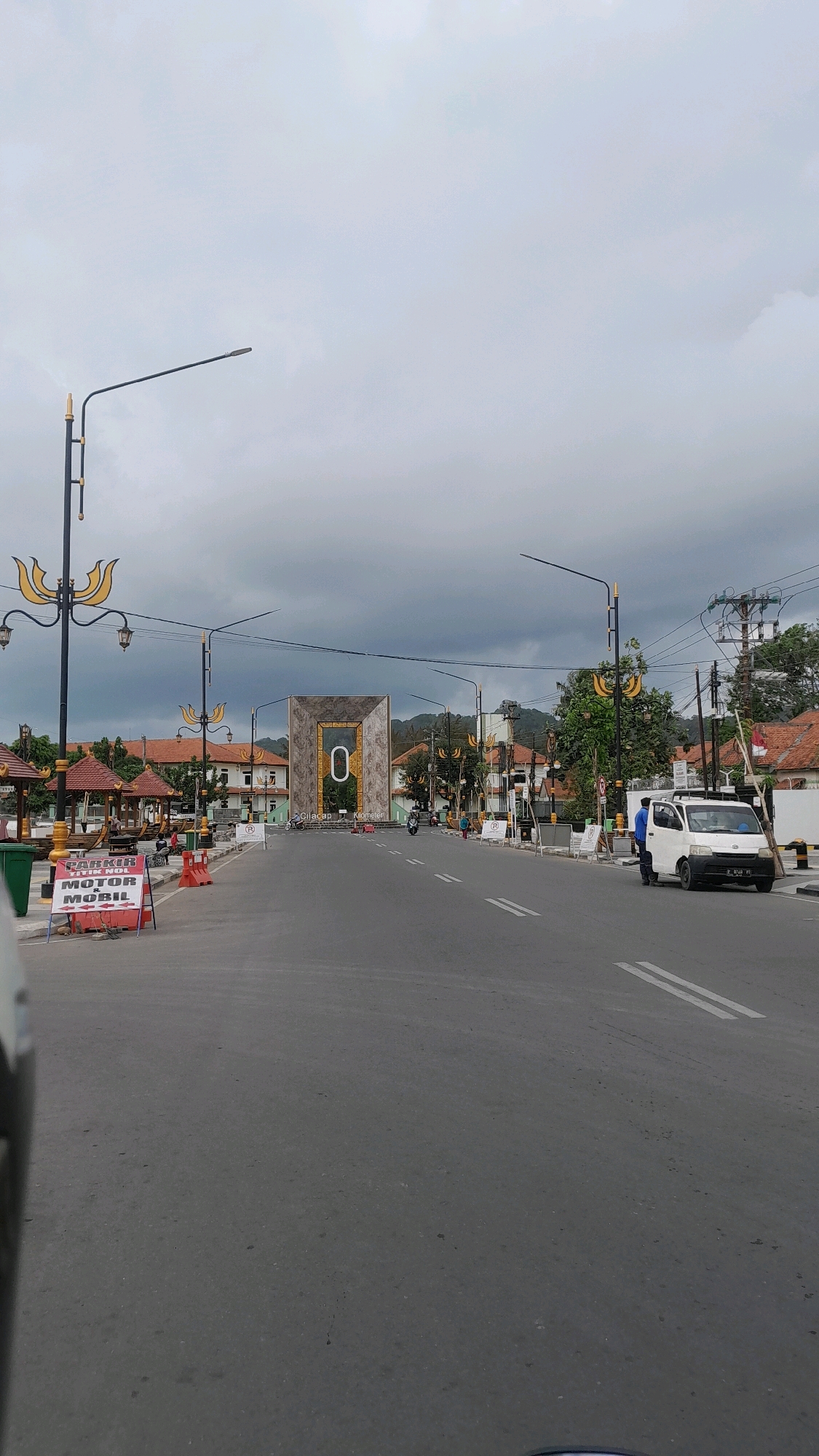 The image shows a wide, paved road in Desa Tambakreja, Indonesia, leading towards a prominent, modern archway featuring a large number "0". The sky is overcast with dark, heavy clouds.
On the left side of the road, a prominent sign reads "PARKING TITIK NOL MOTOR & MOBIL" indicating a parking area for motorcycles and cars at Kilometer Zero. Several people are visible near shaded gazebos and on the left sidewalk, some seated. There are also orange traffic cones on the road and sidewalk.
The archway, a significant landmark, is a large, rectangular structure with decorative elements and the number "0" prominently displayed in its center. The text "Cilacap Kilometer" is visible below the archway.
On the right side of the road, a white van is parked, with two individuals standing near it. A street lamp with ornate yellow detailing stands on the left side of the road. More street lamps with similar designs are visible further down the road. Trees and buildings with red-tiled roofs line the sides of the road. Electrical poles and wires are present in the background, with a hill or mountains visible in the distance. The road is marked with white lines. The perspective of the image suggests it was taken from a moving vehicle.