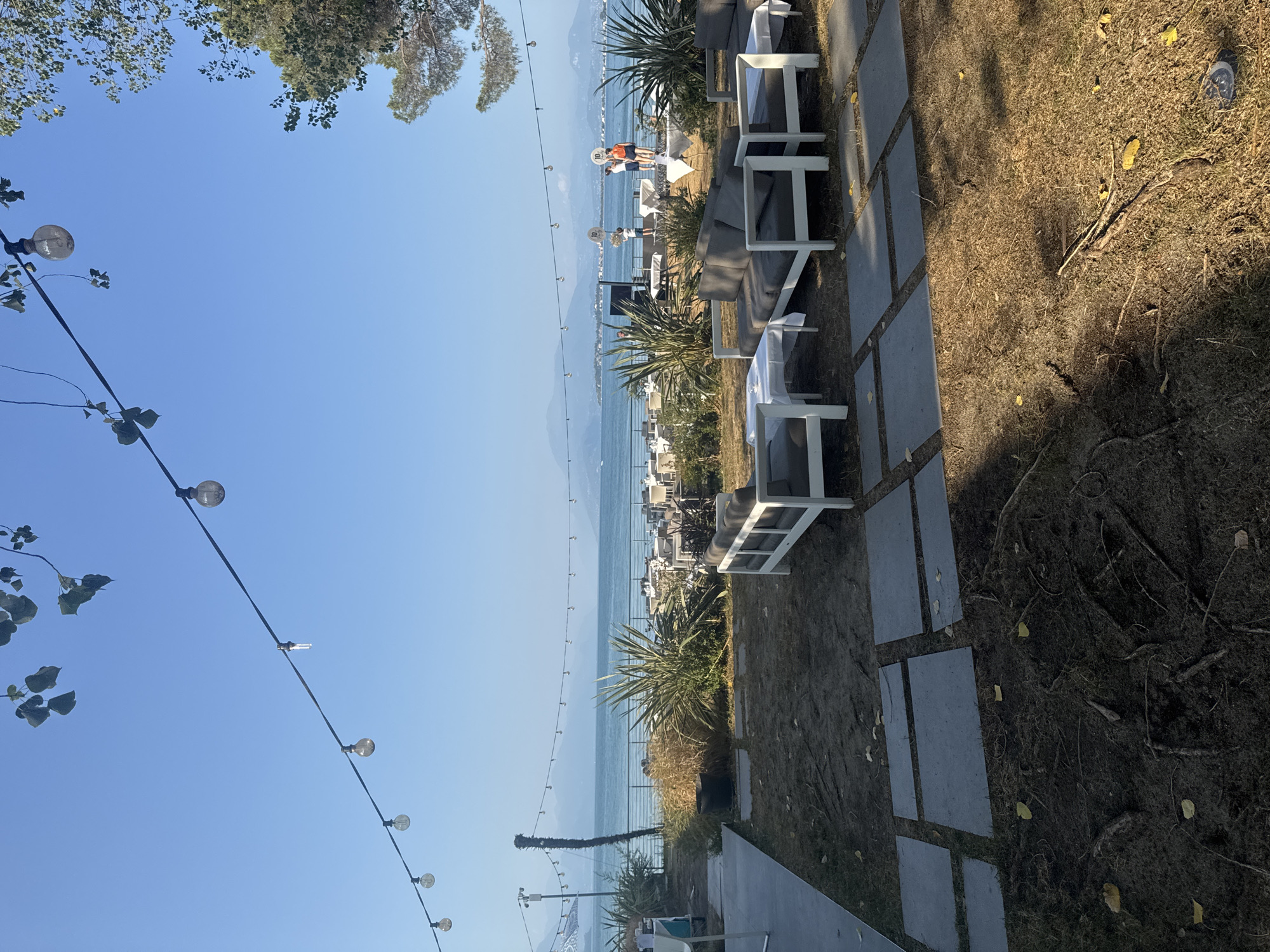 The image depicts an outdoor seating area adjacent to a body of water, likely Lake Garda, in Desenzano del Garda, Italy. In the foreground, there are several low-lying gray sofas with white cushions arranged around white-clothed tables on a patch of grass and soil. Stone pavers are laid out in a path leading towards the seating area. In the midground, a line of lush green bushes and dry grasses separates the seating area from a view of the lake and distant mountains. Beyond the bushes, there are numerous tables and chairs set up, suggesting a restaurant or event space. On a raised platform in the distance, three people are standing, two of whom are holding up signs with the number "10" on them. Overhead, a string of incandescent bulbs hangs across the scene, casting a warm glow. The sky is clear and blue, indicating fair weather.
