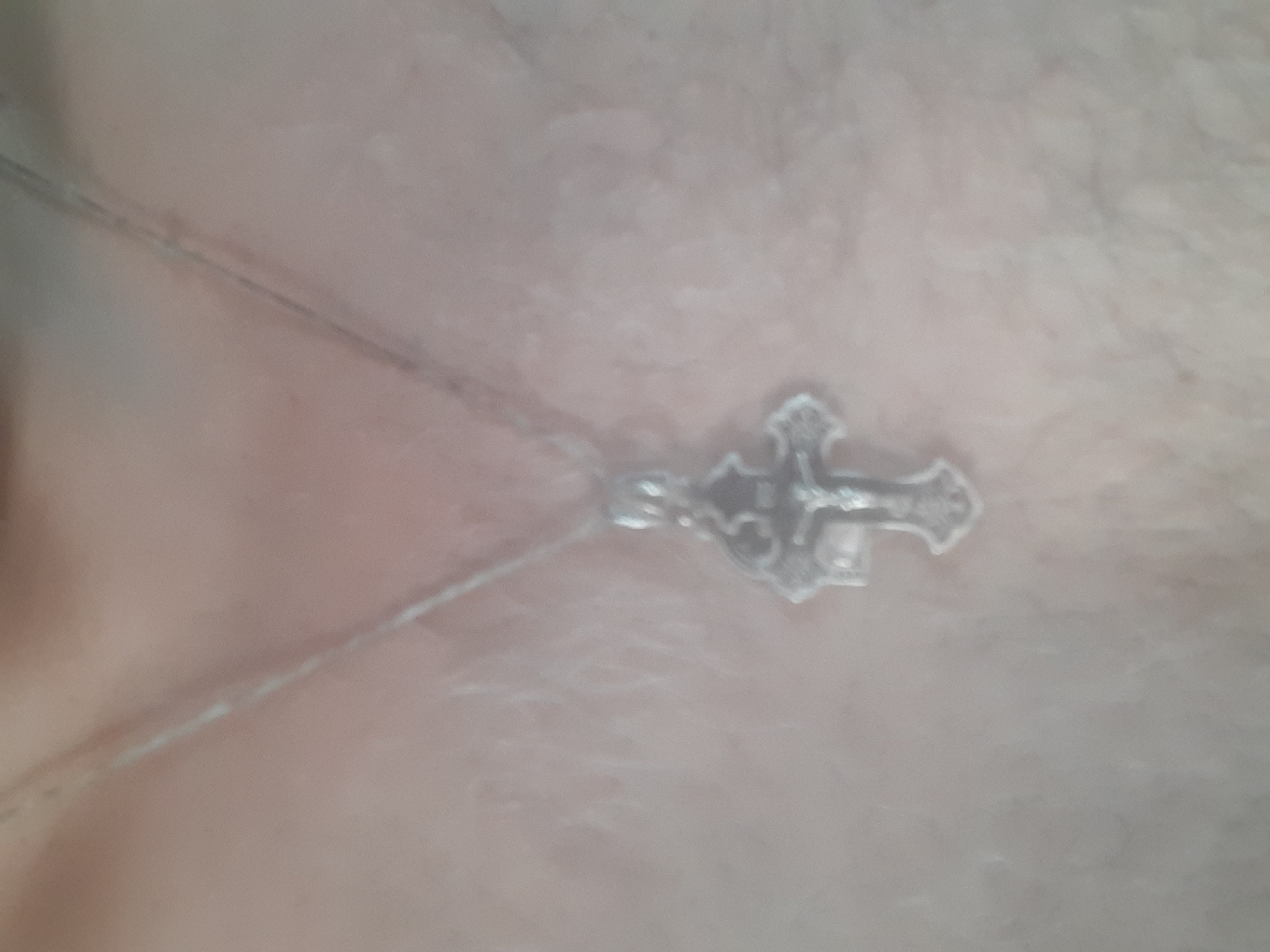 The image shows a close-up of a person's chest or neck with a silver cross pendant on a delicate chain. The pendant is detailed and appears to be made of metal, with intricate designs on its surface. The chain is thin and silver, lying against the skin. The background is softly blurred, suggesting a shallow depth of field, and the skin tone is fair. There are some faint lines or textures visible on the skin, possibly indicating stretch marks or the texture of clothing. The lighting is soft and diffused, creating a gentle illumination on the subject and the jewelry. No text is visible in the image. The overall impression is intimate and personal, focusing on the detail of the cross and its placement on the body. The context of Yessentukskaya, Russia, does not provide specific visual cues in this close-up shot, but it places the image within a particular geographical and cultural setting.