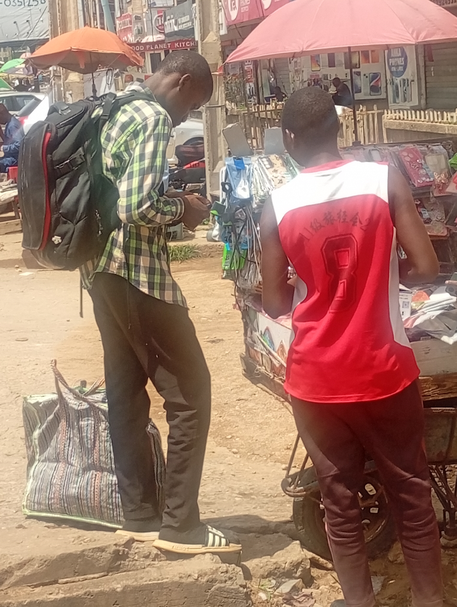 Two men are observed on what appears to be a city street in Maiburiji, Nigeria. The first man, wearing a green plaid shirt, black pants, and black and white Adidas-branded sandals, has a large black backpack and a patterned tote bag. He is looking down at something in his hands, possibly a mobile phone. Beside him, another man wearing a red and white jersey with the number "8" on the back, paired with brown pants, is standing at what appears to be a vendor's cart. The surroundings include other vendors' stalls under colorful umbrellas, various signage in the background, and parked cars. The setting suggests a bustling street market or shopping area.