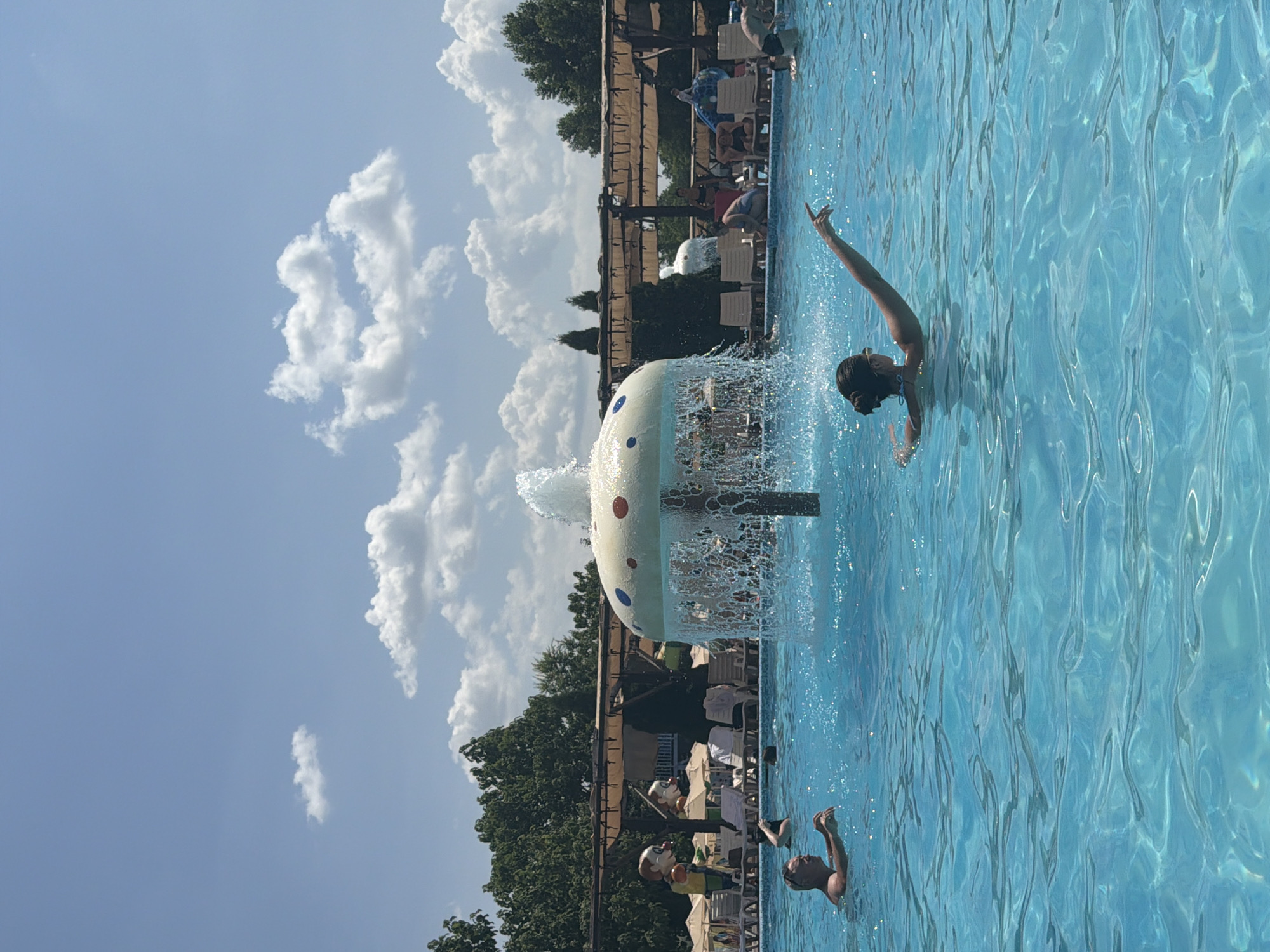 In the city of Sociteni, Moldova, a clear blue swimming pool is teeming with activity on a sunny day. The water is disturbed by ripples and splashes, indicating movement. In the foreground, a woman with dark hair pulled back in a bun is swimming, her left arm extended upwards and pointing towards the right. To her left and slightly further back, a man with short, gray hair and sunglasses is partially submerged in the water, his arms bent forward as if reaching out or treading water.
Further back in the pool, other individuals can be seen, though their features are less distinct. A mushroom-shaped fountain is centrally located, spraying water upwards, creating a misty cascade. In the background, the poolside is lined with lounge chairs and umbrellas, some occupied by people. Beyond the pool area, a line of trees forms a natural backdrop, and the sky above is a bright blue with scattered white clouds. Large, cartoon-like figures of clowns with yellow hair and red noses are visible above the lounge chairs, adding a playful element to the scene.