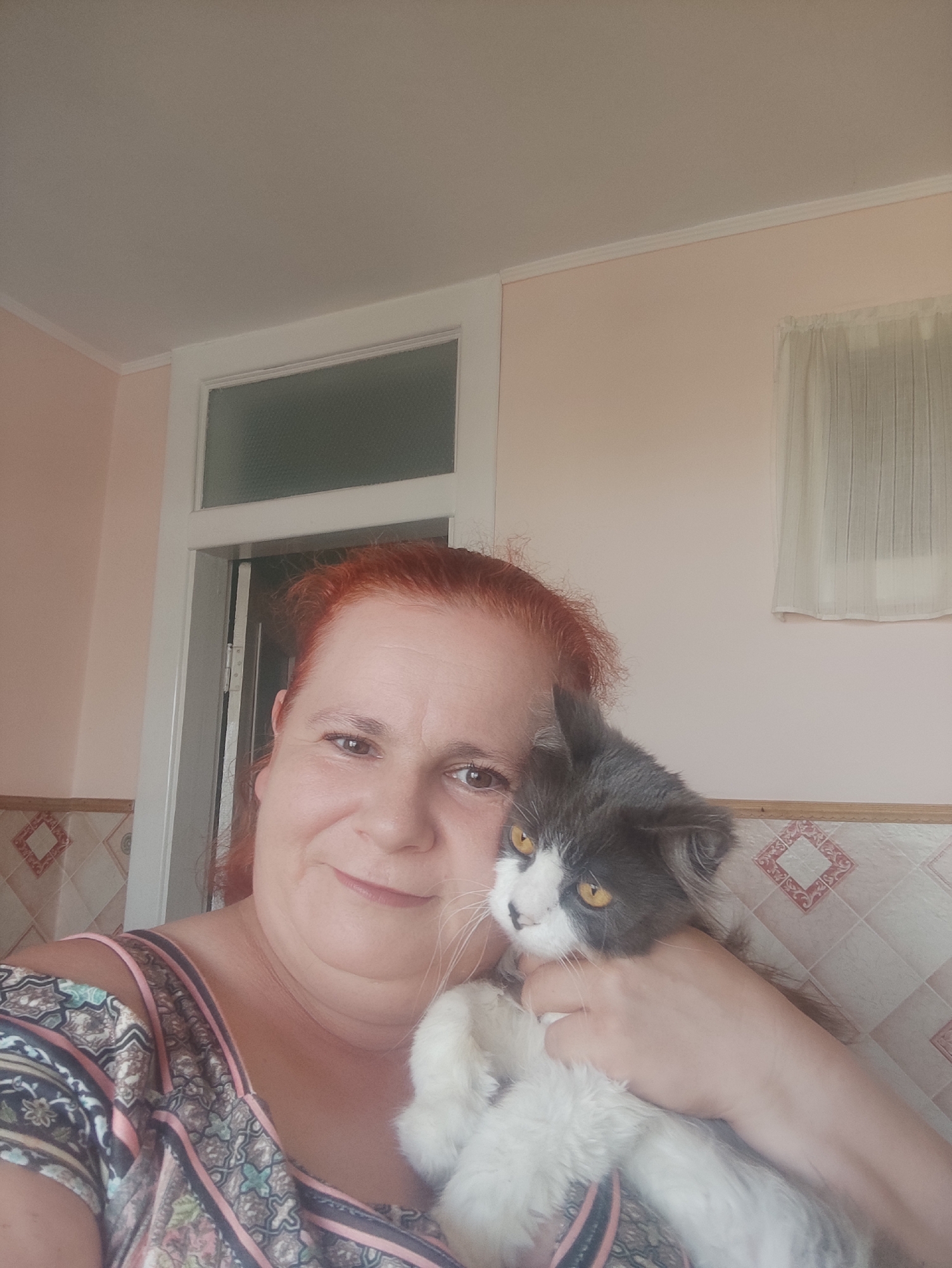 A woman with red hair is taking a selfie, holding a gray and white cat. The woman is smiling and looking at the camera. The cat is looking slightly to the side with yellow eyes. They are indoors, in what appears to be a room with light pink walls. There is a window with a white curtain in the background and a doorframe visible on the left side of the image. The woman is wearing a colorful patterned top. The overall impression is a casual and affectionate moment. There is no visible text in the image.