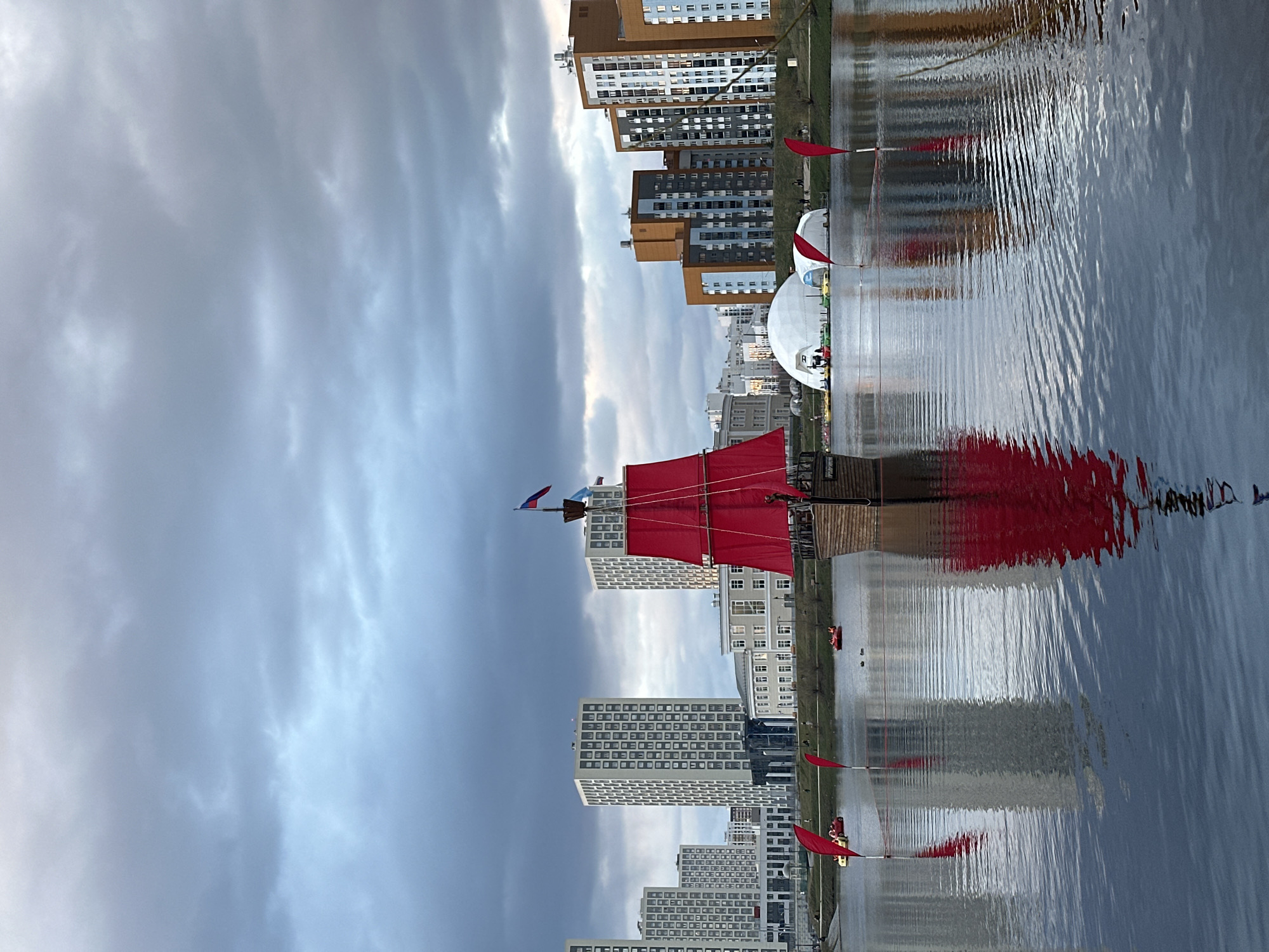 The image shows a ship with red sails on a body of water in Yekaterinburg, Russia. The ship is sailing from left to right. Modern buildings are visible along the shoreline, and the sky is cloudy. There are also white, dome-shaped structures located alongside the water. A series of red flags are spaced across the surface of the water.