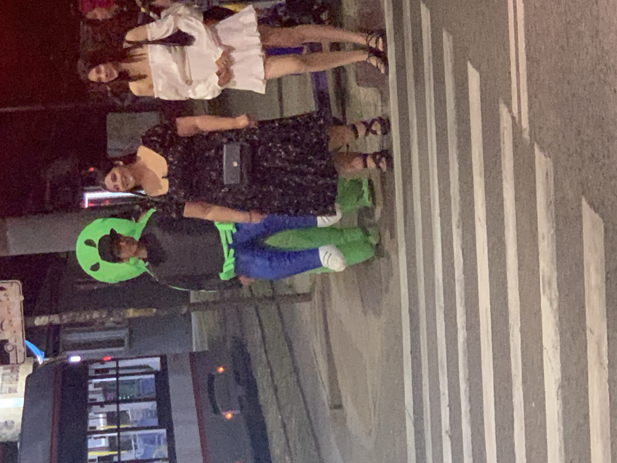 In an urban setting at night, three individuals stand on a pedestrian crossing. On the left, a person in a bright green alien costume with a black cap and dark clothing, including blue pants, faces towards the right. The costume appears to be inflatable. To the right of the alien, a woman in a dark, floral-patterned dress with puffy sleeves and dark sandals smiles as she looks to her left. Next to her, a woman in a white off-the-shoulder dress and black sandals looks forward. A black purse is slung across the first woman's chest. A tram is visible in the background on the left, with its lights illuminated. The scene is dimly lit, indicating it is nighttime.
