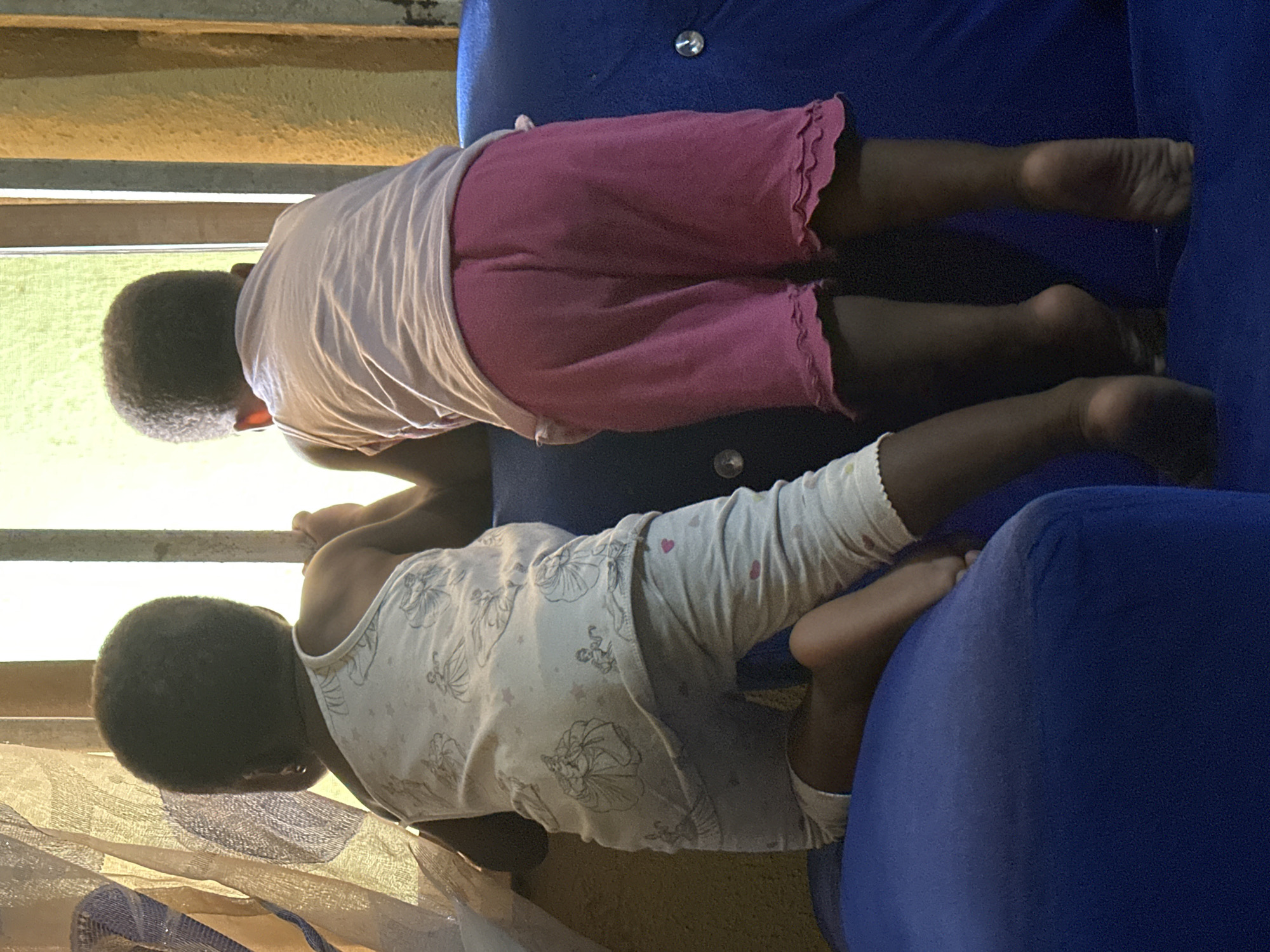 In Narayi, Nigeria, two children, seen from behind, stand on a blue sofa, peering out a window. The child on the left wears a white tank top with an illustration of princesses and light gray pajama bottoms. The child on the right wears a pink t-shirt and pink ruffled shorts. A sheer curtain hangs to the left of the window, and the wall to the right is textured and yellow. The sunlight streaming through the window creates a bright, almost silhouetted effect on the children.