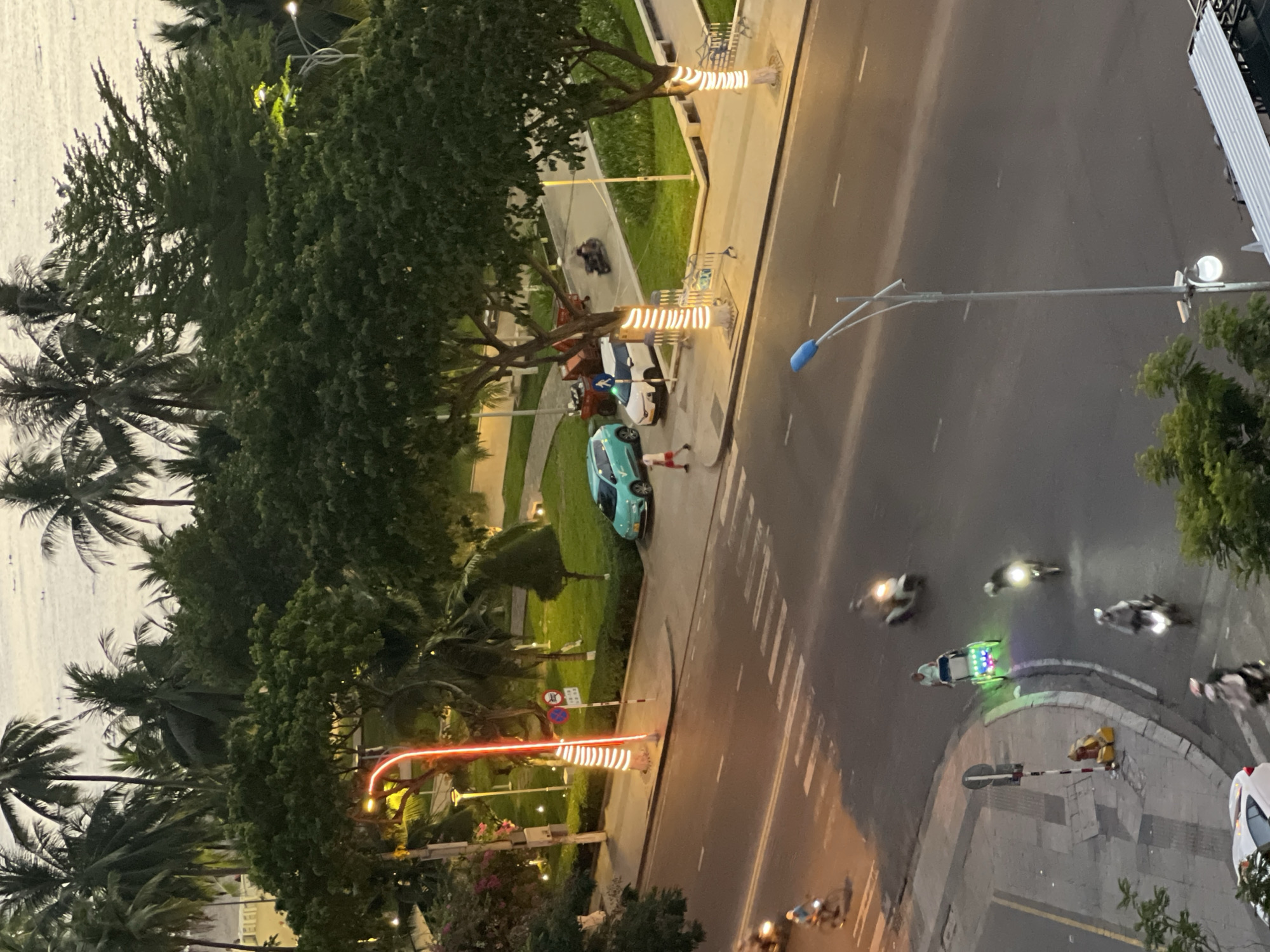 This is a high-angle, long shot of a street scene in Nha Trang, Vietnam, likely during dusk or early evening. The foreground shows a road with several motorbikes and a cyclos being ridden, some with illuminated lights. One cyclos is particularly noticeable with its bright green and colorful LED lights. A pedestrian wearing a white shirt and red shorts is walking across a crosswalk. On the side of the road, there are a few parked cars, including a teal-colored vehicle and a white one. The background features lush green trees, palm trees, and a glimpse of the ocean or a large body of water. Streetlights and decorative lights wrapped around tree trunks are visible, illuminating the scene. There are also signs and benches along the sidewalk. The overall impression is a bustling urban environment with tropical foliage and coastal proximity.