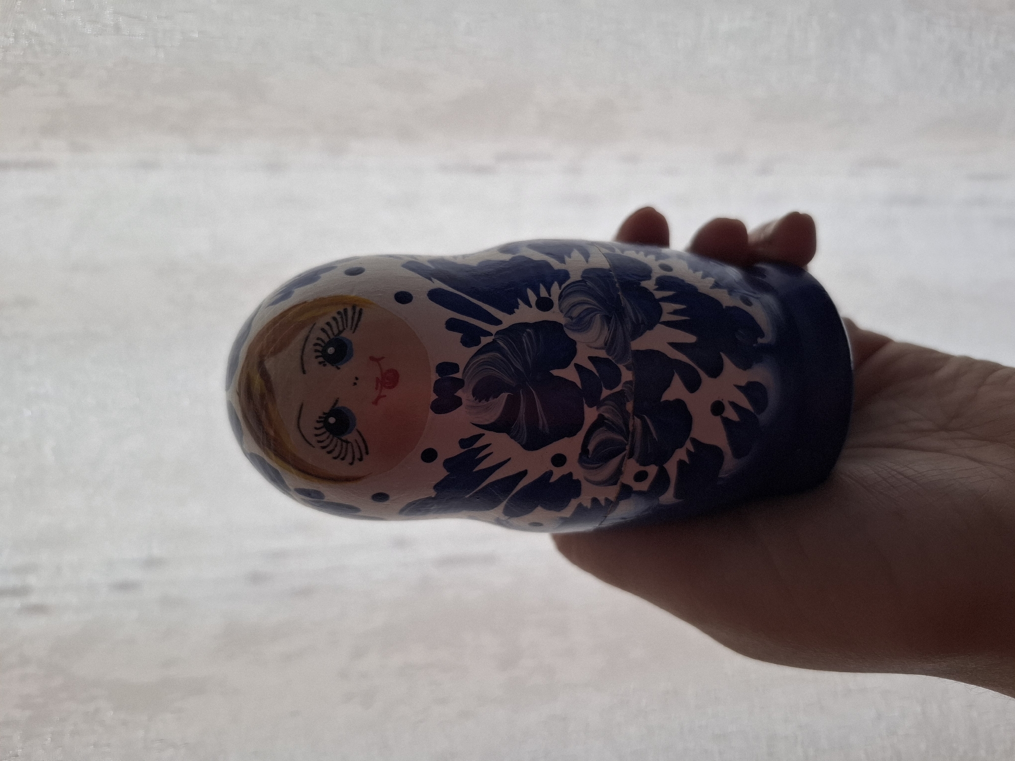 A hand holds a Russian nesting doll against a bright, neutral background. The doll is painted with a face and floral designs in dark blue. The city location is not discernible from the image.