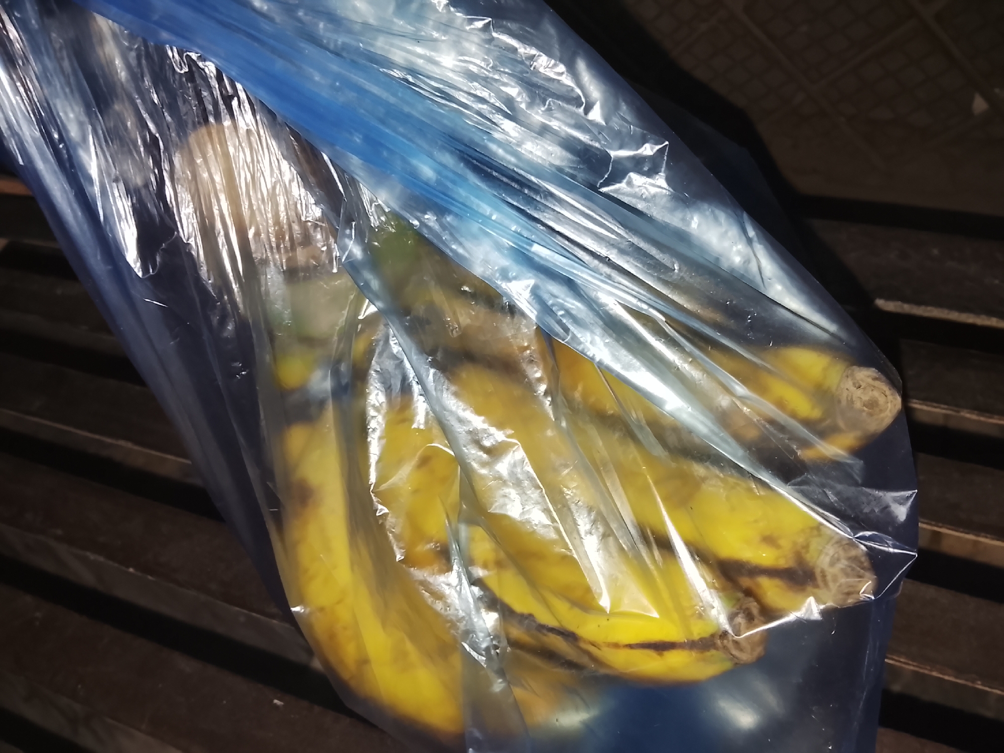 A close-up shot features a bunch of ripe, yellow bananas contained within a translucent, blue-tinted plastic bag. The bananas show signs of ripeness with some brown spots on their skins. The plastic bag is visibly crinkled, reflecting light and creating highlights and shadows on its surface. The background is dark and indistinct, appearing to be a wooden or slatted surface with visible horizontal lines, possibly a shelf or a crate. There is no specific activity depicted, as the bananas are stationary in the bag. The lighting is soft and diffused, suggesting an indoor setting or a shaded area. No people are visible, and there is no visible text in the image. The scene is observational, focusing on the everyday objects.