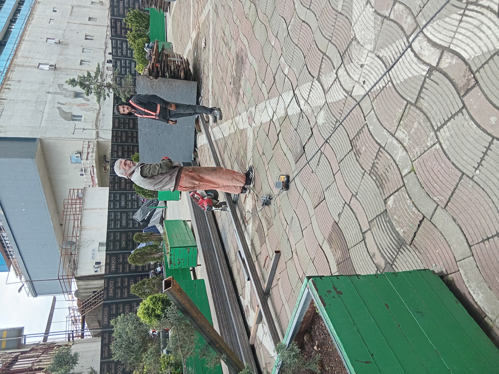 The image captures a street scene in Batumi, Georgia, where two men appear to be engaged in metalworking. The location seems to be a public pedestrian area due to the presence of patterned paving stones, potted plants, and benches. One man, older with white hair, is positioned next to metal rods and a red angle grinder, presumably cutting or preparing the metal. The second man stands further off, watching or assisting. The environment includes a building with multiple stories covered with "COCHEZ" marked construction sheets. There is also a metal structure above a building staircase. The weather appears to be sunny, casting shadows in the scene.