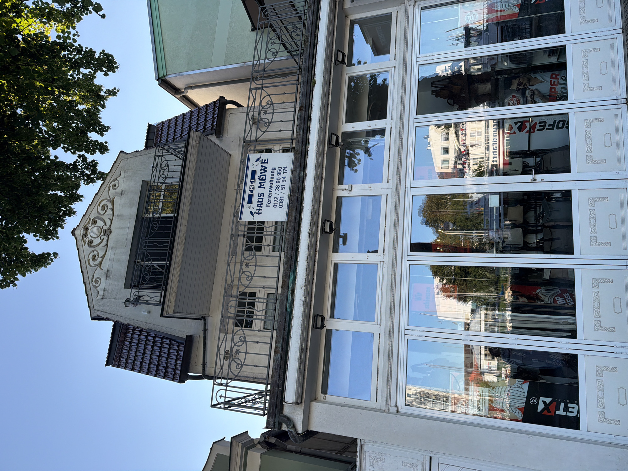The image shows the facade of a building in Rostock, Germany, with a clear blue sky above. The building has multiple levels, with a prominent balcony on the upper floor featuring decorative iron railings. A sign on the balcony reads "FREI HAUS MÖWE Ferienwohnung" (Free Seagull House Holiday Apartment) followed by phone numbers. The lower part of the building features a storefront with large glass windows and doors, reflecting the surrounding environment. The storefront displays merchandise and advertising posters, including one that reads "SUPER SALE". The architectural style appears to be a mix of traditional and more modern elements. Trees and other buildings are visible in the reflection of the storefront windows, suggesting an urban setting near a waterfront or harbor.