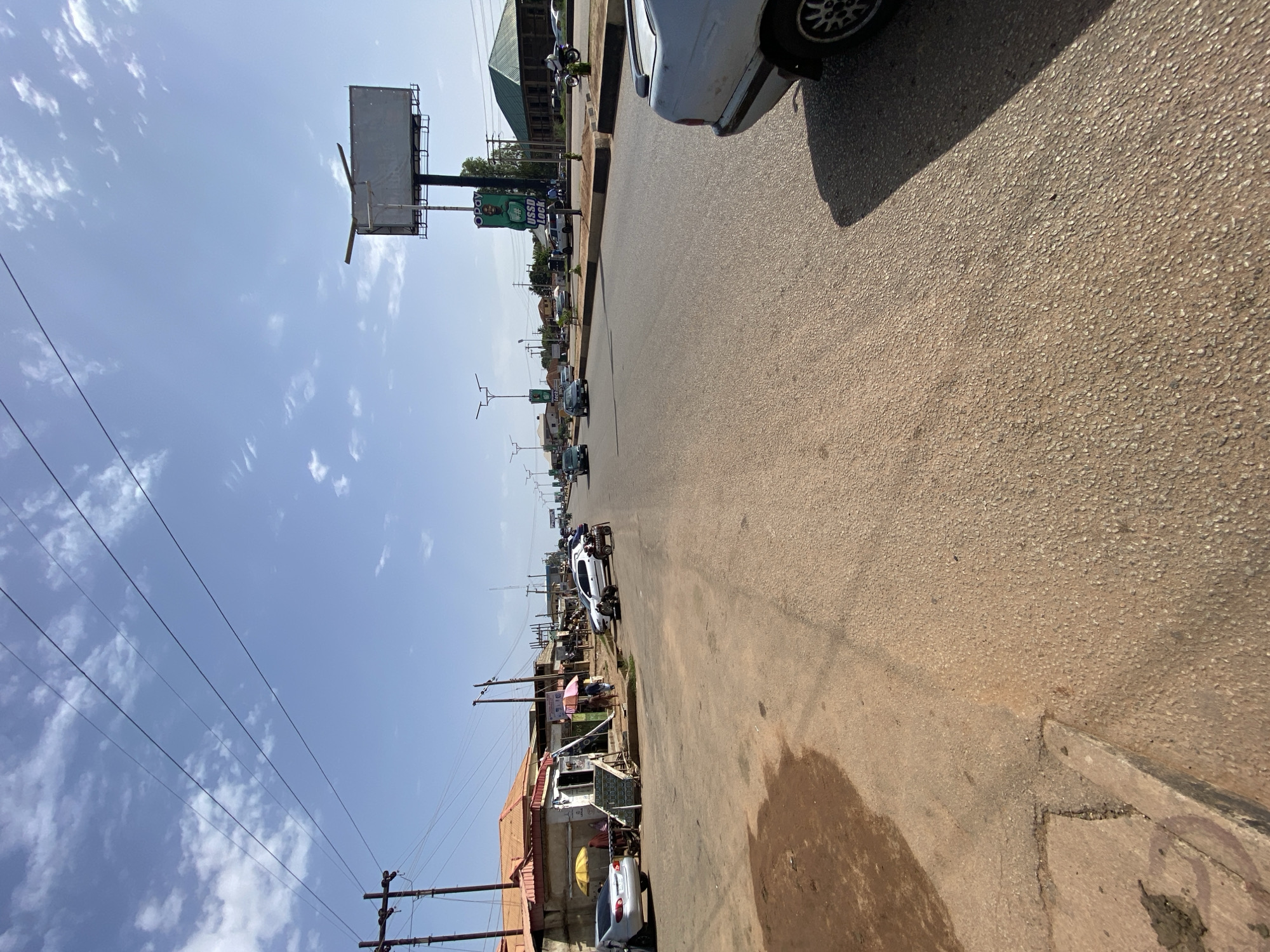 The image depicts a street scene in Gura Top, Nigeria, under a clear blue sky with scattered white clouds. The road is paved and appears well-trafficked, with several vehicles visible, including cars and a motorcycle. On the left side of the road, there are buildings with orange roofs and a small market stall with people gathered. A large billboard stands on the right side of the road, displaying advertisements. Utility poles and power lines are visible throughout the scene, typical of urban infrastructure. The overall impression is of a bustling commercial area on a sunny day.