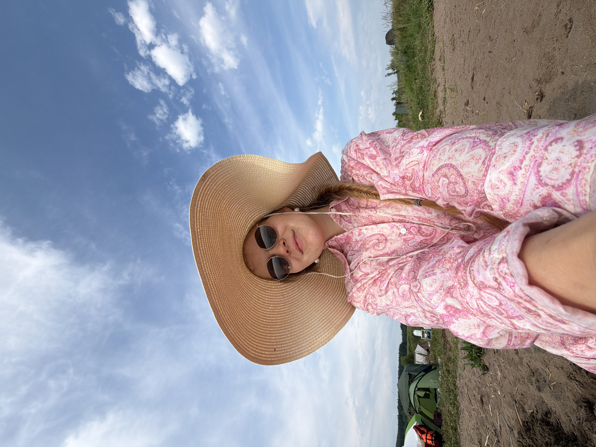 A woman wearing a large straw hat and a pink paisley shirt is photographed from the chest up. She is wearing round sunglasses and has a blonde braid. The background shows a beach with tents and vegetation under a cloudy blue sky. The image appears to be taken in the city of Krasny Bor, Russia, likely near the Neva River, given the sandy terrain and presence of tents which suggest a recreational area or campsite.