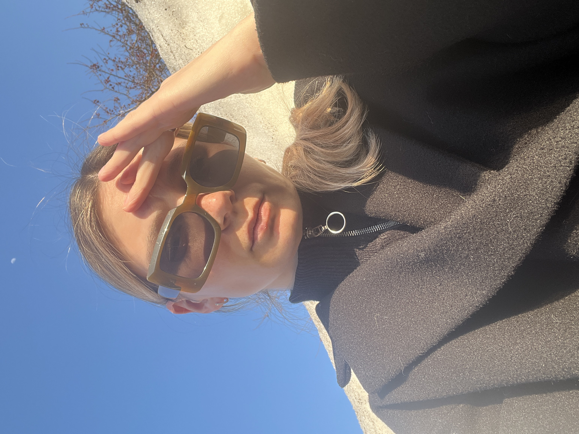 A partial portrait-style image, likely a selfie, depicts a light-skinned female individual from the chest up, under strong natural illumination. The subject is wearing large, olive-green rectangular sunglasses, a black high-collared garment with a silver circular zipper pull and zipper, and a dark, textured overcoat. Her right hand is raised, fingers splayed, partially covering her forehead and the top edge of her sunglasses, consistent with shielding her eyes from direct sunlight. Blonde hair is visible around her face and as a tied-back portion over her left shoulder. A small silver hoop earring is present in her left ear. The background consists of a clear, bright blue sky, with a faint crescent moon visible in the upper left quadrant. To the right and behind the subject, a bright, irregular, textured mass, possibly a snowdrift or light-colored ground feature, is present, with bare tree branches featuring small red berries emerging above it. The precise location within the city of Chaikovsky, Russia, is not discernible from the image content.
