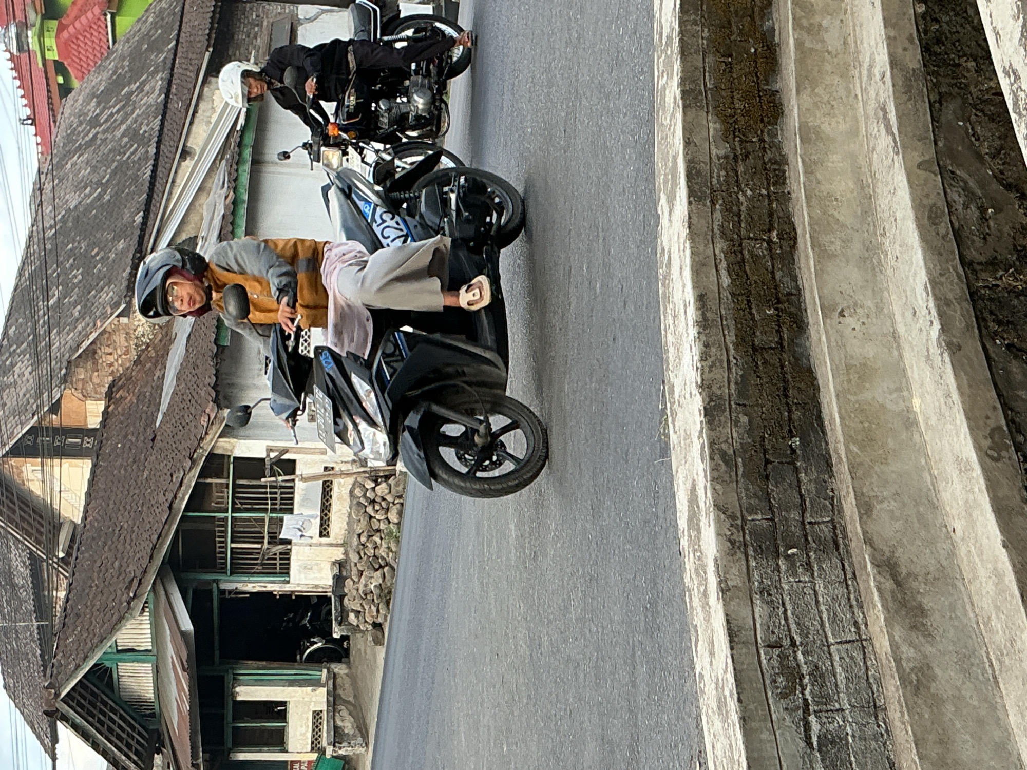In the city of Kebonrejo, Indonesia, two individuals are riding motorcycles on a paved road. The person in the foreground is wearing a helmet and a tan jacket over a light pink garment, riding a black scooter with blue markings. They are positioned on the left side of the road, with their legs visible. Behind them, another person wearing a white helmet and a dark jacket rides a black motorcycle. The background features traditional Indonesian houses with tiled roofs and stone walls, suggesting a residential or commercial area within the city. The road surface is asphalt, and a concrete structure is visible in the foreground, possibly a drainage system or a raised curb. The scene appears to be during daylight.