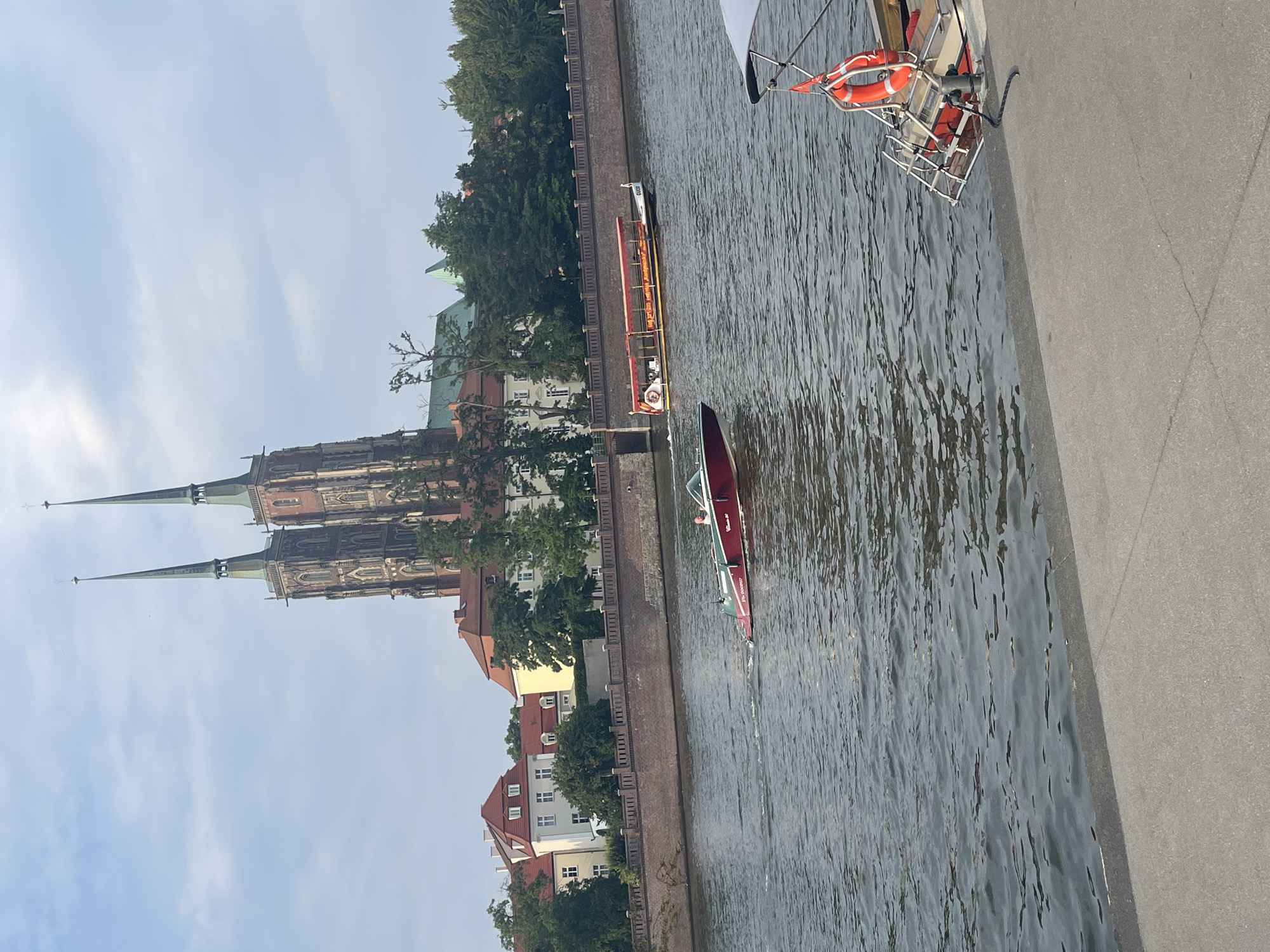 In Wrocław, Poland, the image captures a section of the Oder River with a prominent view of the Cathedral of St. John the Baptist in the background. The Gothic cathedral features two tall spires and is partially obscured by trees and other buildings. In the foreground, two boats are visible on the water. One is a smaller, red motorboat with a person at the helm, creating a wake. The other is a larger, yellow tour boat with a red canopy, carrying several passengers. To the right of the frame, a portion of another boat with a white canopy, a life preserver, and mooring rope is visible at the edge of the water, adjacent to a concrete embankment. The sky is partly cloudy, indicating daytime.