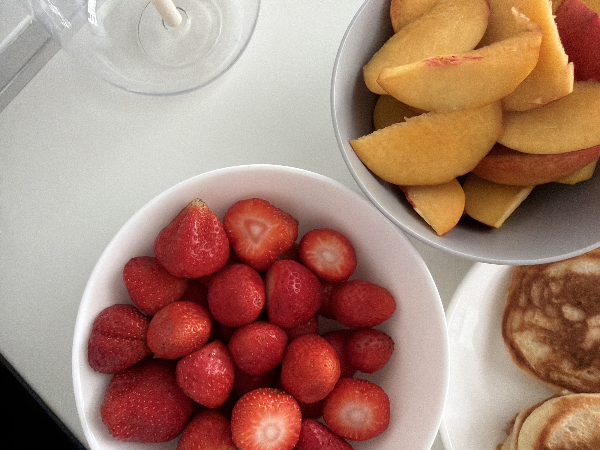 A top-down, close-up shot features a breakfast spread on a white surface, likely in Finestrat, Spain. In the foreground, a white bowl is filled with sliced peaches. To the left, a white bowl overflows with whole and halved strawberries. Below the strawberries, a white plate holds two golden-brown pancakes. In the upper right corner, a clear glass with a straw is partially visible. The scene is well-lit, highlighting the vibrant colors of the fruit.