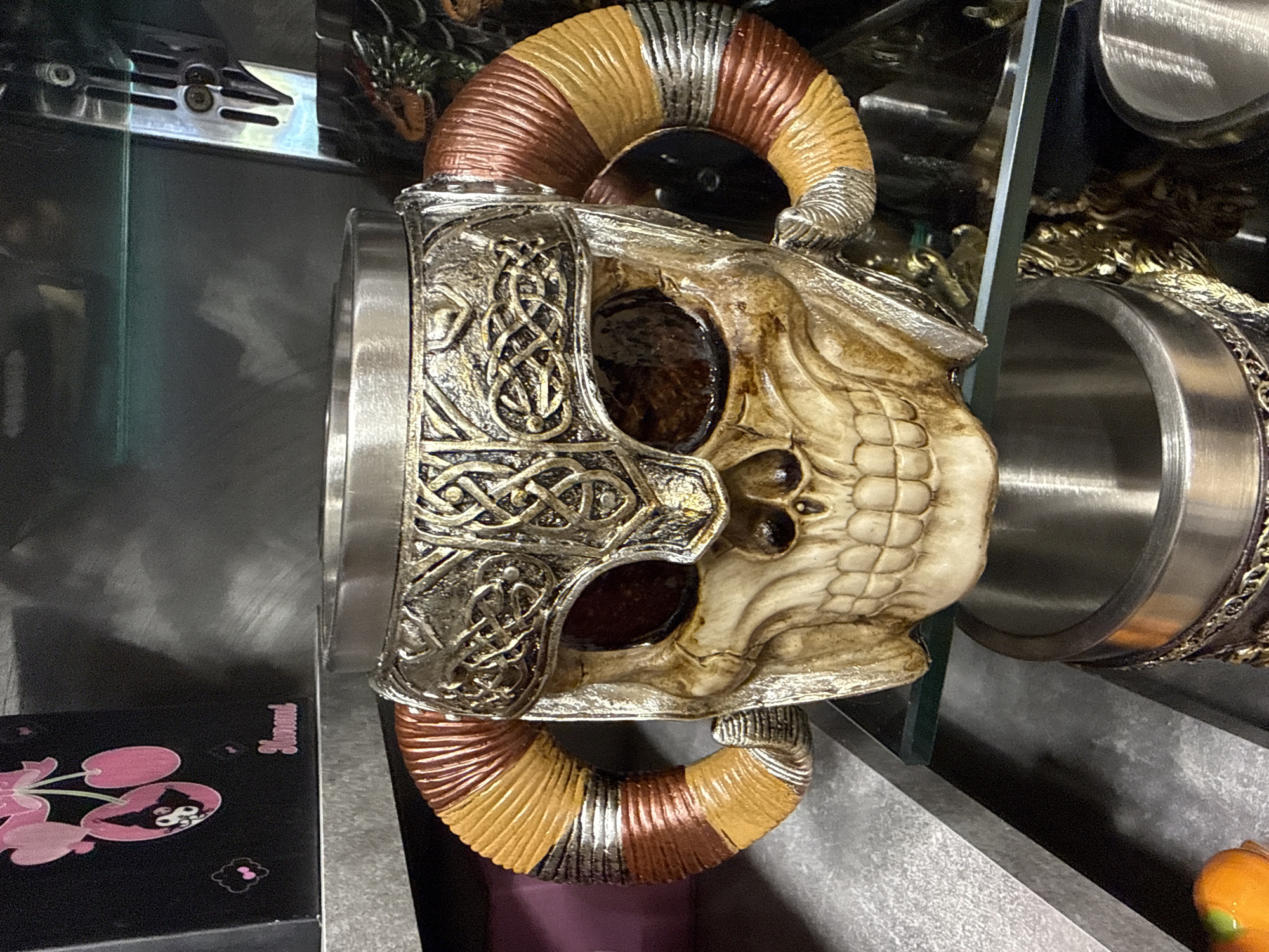 The image depicts a detailed, decorative skull-themed mug or tankard displayed on a clear glass shelf within an interior setting, likely a retail store or collection cabinet. The central object is a large, ornate vessel featuring a skull facade with dark, hollow eye sockets and visible teeth. The top of the skull is adorned with a metallic-finish headpiece, intricately detailed with interwoven Celtic knot patterns, appearing to be made of an aged silver-bronze material. Two curved, ribbed horns extend from the sides, banded in alternating shades of orange, yellow, brown, and silver. A smooth, reflective metallic rim is visible at the top opening of the vessel.
Below the clear glass shelf, the top portion of another similar metallic-lined vessel is partially visible. To the upper left of the main subject, a black rectangular box is present on a higher, textured grey shelf. This box features pink graphics, specifically two cherries with stylized skull faces, and the word "Kuromi" printed in pink script. To the upper right, a metallic, vertically oriented fixture or bracket is visible against a dark background, showing screw details. Further into the background, behind the main skull mug, partially obscured ornate objects with hints of green and orange can be discerned, possibly another decorative item with a dragon motif. The overall environment suggests a display or shelving unit with artificial lighting. There are no people or specific actions visible in the scene. The location is in the city of Primorsko, Bulgaria.