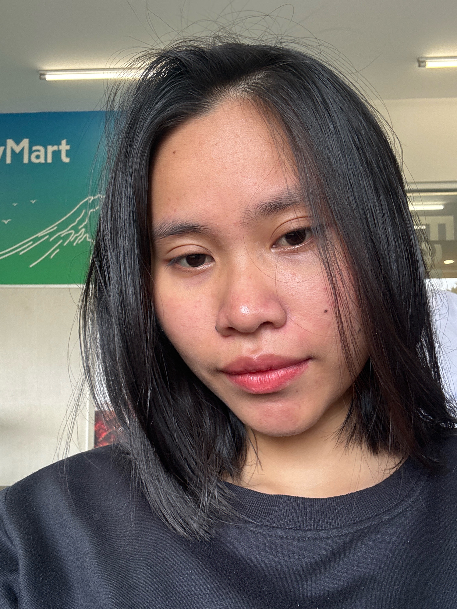 A young woman with dark hair and medium-toned skin is shown in a close-up, waist-up portrait. She is wearing a black crewneck sweatshirt and is positioned in front of a wall with a mural depicting mountains and birds. A fluorescent light fixture is visible on the ceiling above. The background suggests an indoor location, possibly a convenience store like "FamilyMart," given the branding visible on the mural. The woman's face is slightly tilted, and she is looking downwards. Her expression is neutral, with her lips slightly parted. She has a mole on her left cheek, and her skin shows some texture and mild redness.