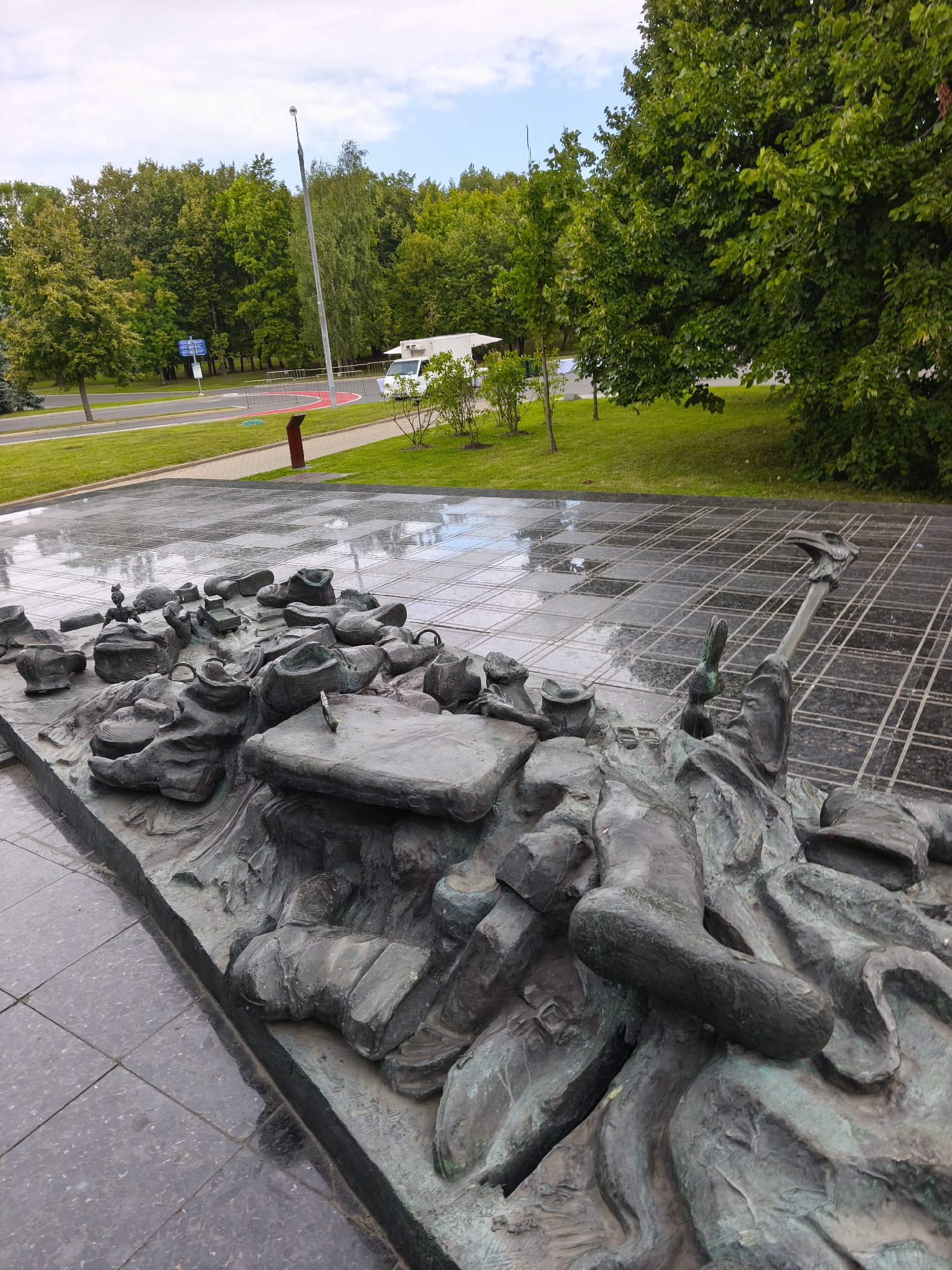 A bronze sculpture depicting a chaotic scene of figures and objects is situated on a wet, tiled plaza. The sculpture appears to be a relief, with various elements raised from a base. Among the discernible forms are what seem to be shoes, fragments of bodies, and abstract shapes, all rendered in a dark, aged bronze with hints of green patina. The plaza itself is paved with large, dark gray tiles, many of which are wet and reflecting the overcast sky. In the background, a lush green park with numerous trees dominates the landscape. Beyond the park, a street is visible, with a white van parked on the side and a lamppost rising towards the sky. The overall impression is that of an outdoor art installation within a park or public space. This is the monument to the Victims of the Kleshchev District, Moscow, Russia.
