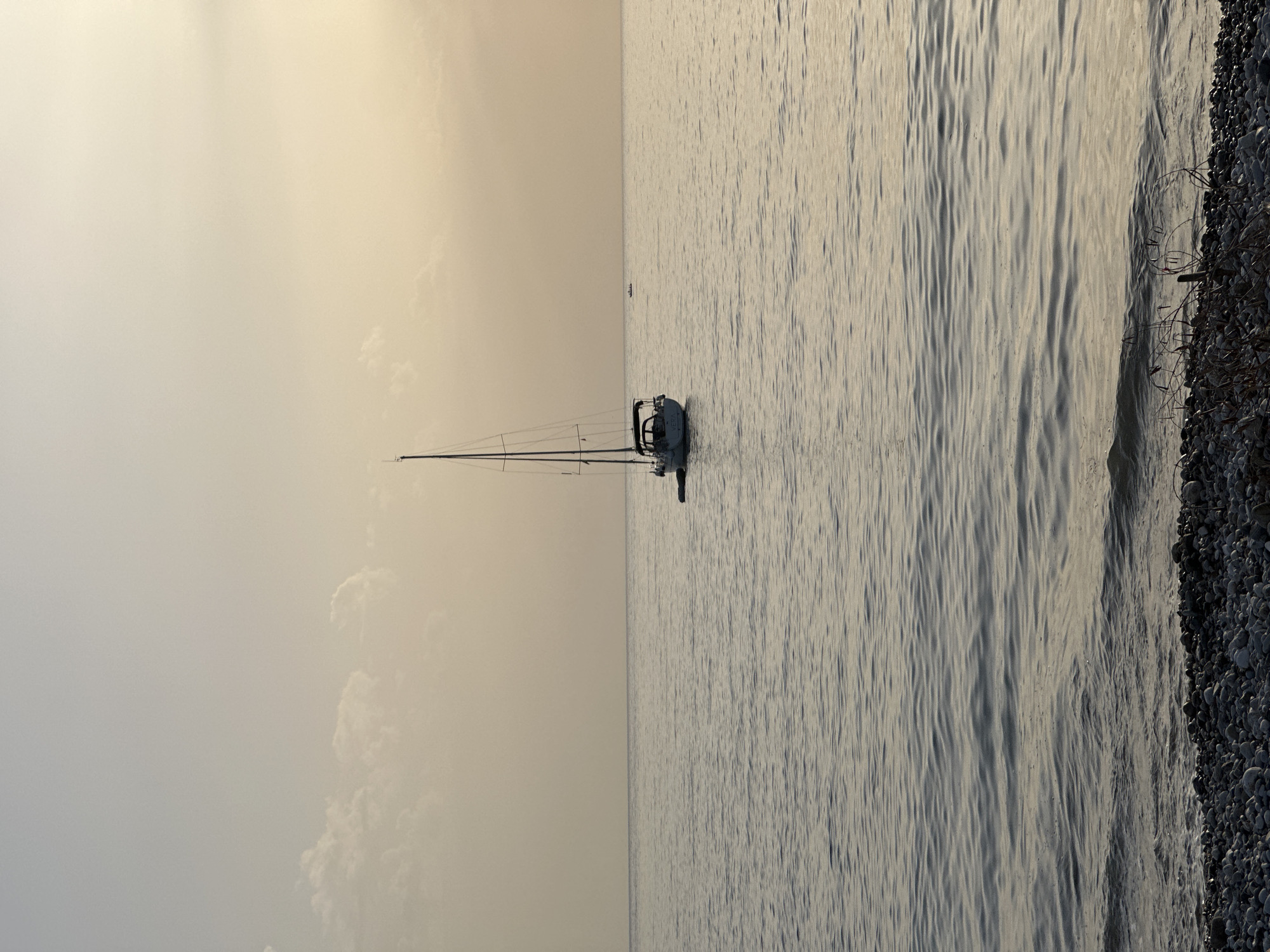 The image shows a sailboat anchored off the coast of Šušanj, Montenegro. The sailboat is white with a dark mast and rigging. The sea is calm with small waves, and the sky is overcast with a light, pale color. A rocky shoreline is visible on the right side of the image. In the distance, another boat can be seen.