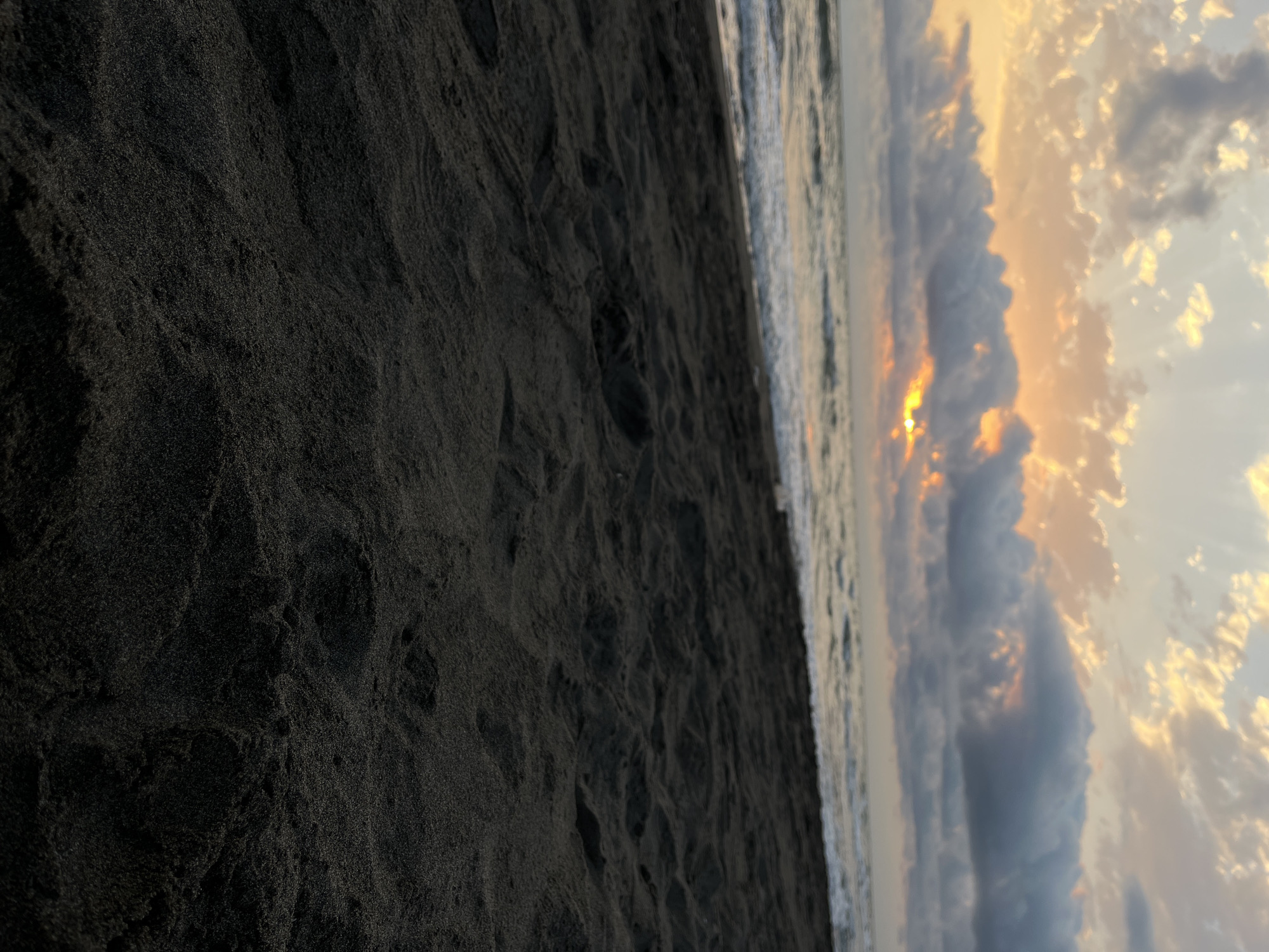 The image captures a serene beach scene at dusk. The foreground is dominated by dark, granular sand, suggesting it might be volcanic sand common in some coastal areas of Georgia. The sand exhibits a textured surface with subtle undulations, hinting at wind or water movement.
In the midground, the gentle waves of the Black Sea break onto the shore, creating a foamy white line. The water appears calm with a slight ripple.
The background is a dramatic sky filled with clouds. The sun is setting, peeking through gaps in the clouds, casting a warm, orange glow. Shafts of sunlight pierce through the cloud cover, creating a beautiful, atmospheric effect. The clouds themselves are a mix of dark, heavy formations and lighter, wispy ones tinged with the colors of sunset.
The location is identified as the community of Supsa in Georgia. This implies the image was taken on the Black Sea coast in that region. There are no people or man-made objects visible in the frame, focusing solely on the natural elements of the beach, sea, and sky.