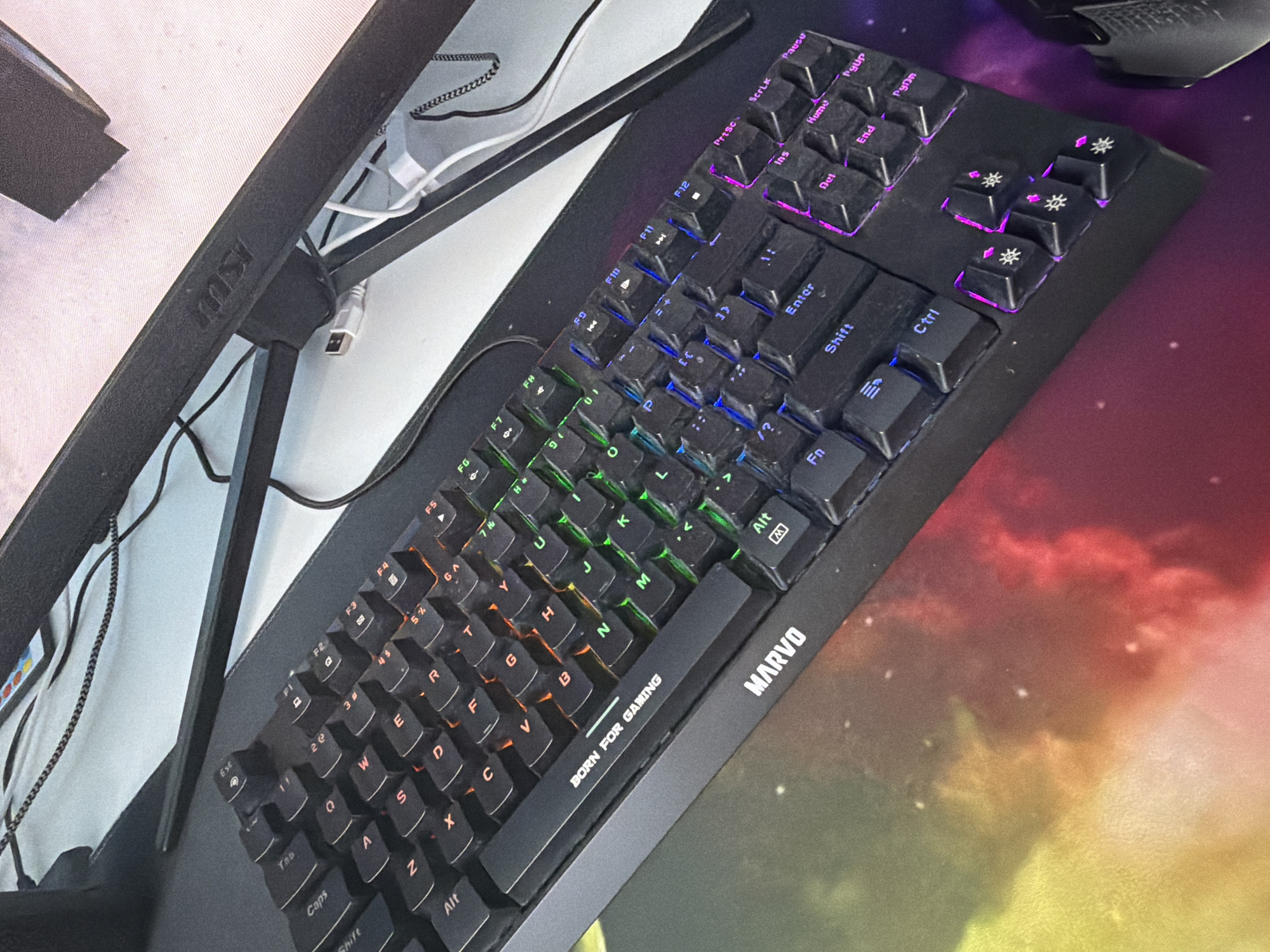 A medium shot depicts a partially visible computer monitor on the left side, its frame inscribed with the word "MSI". To its right, a black "Marvo" keyboard angles upwards from the lower-left to the upper-right of the frame. The keyboard is backlit with varying colors. Below the keyboard, a colorful mouse pad features a blend of red, yellow, green, and purple hues. The overall setting appears to be an indoor gaming or work environment.