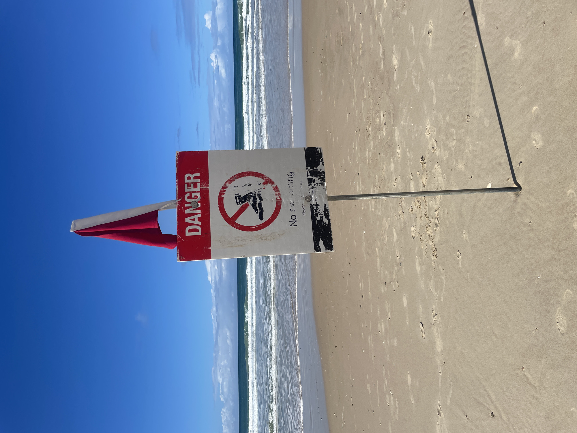 A "DANGER NO SWIMMING" sign with a red and white flag is posted on a sandy beach in the city of Gold Coast, Australia. The sign is weathered and shows some wear and tear. A red circle with a diagonal line across a silhouette of a swimmer indicates the prohibition. The ocean with moderate waves is visible in the background, and the sky is blue with scattered clouds. Footprints are present in the sand, indicating previous activity. The sign is mounted on a metal pole that casts a shadow on the sand.