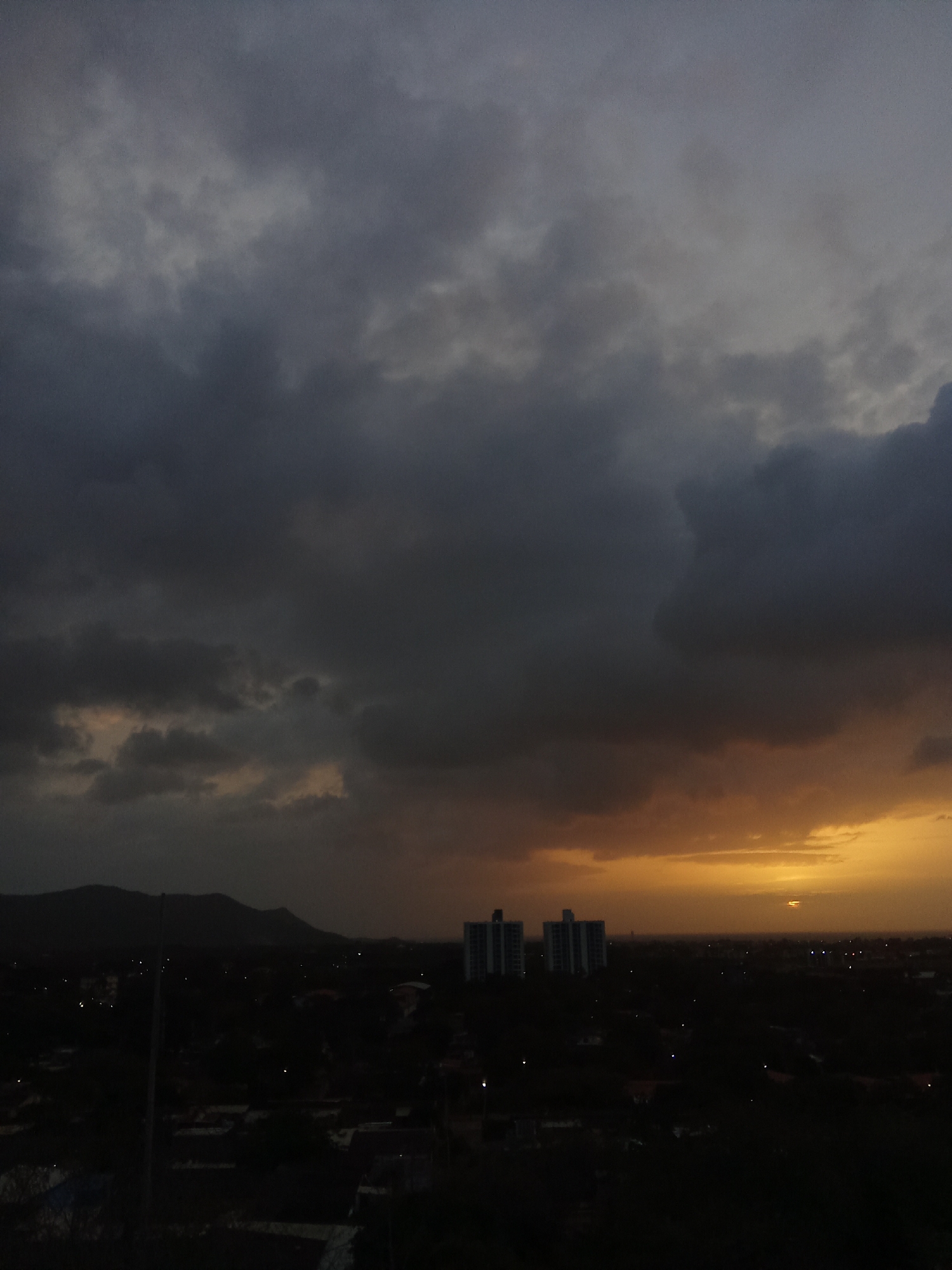 The image depicts a dramatic cloudy sky over the city of Juan Griego, Venezuela, at dusk. Dark, heavy clouds dominate the upper portion of the frame, with hints of orange and yellow light breaking through on the horizon. This suggests an approaching storm or the aftermath of one. In the mid-ground, two tall, modern buildings stand out against the darkening landscape, likely apartments or offices. Below these, the city is largely obscured by shadows, with scattered lights indicating residential or commercial activity. A range of hills or mountains forms a silhouette in the far distance, adding depth to the scene. The overall impression is one of impending weather change and the quiet stillness of a city at twilight.