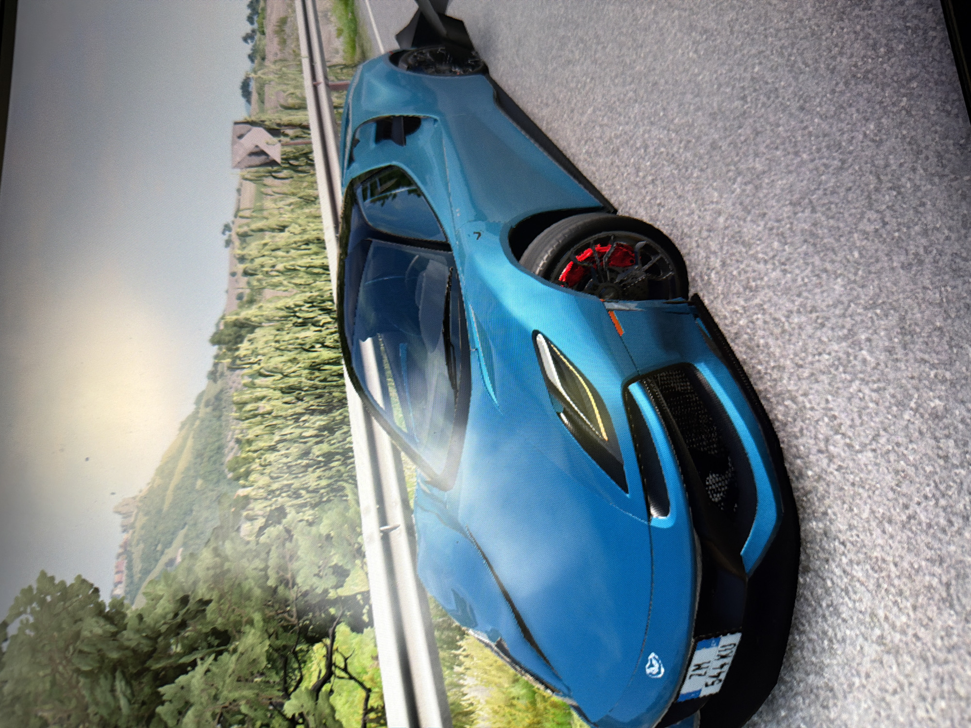 The image displays a sleek, bright blue sports car positioned on an asphalt road. The car is the primary subject, featuring a modern, aerodynamic design with black accents, including a prominent rear wing, front grille, and side skirts. Red brake calipers are visible through the spokes of the front wheel, adding a contrasting detail. A small, indistinct emblem is present on the front hood, and the number "7" is subtly placed on the car's side, near the rear window.
The car appears stationary, centered on the road. The background reveals a verdant, hilly or mountainous landscape. Lush green trees, bushes, and vines cover the hillsides, suggesting a natural, possibly rural or remote setting. A faint, light-colored structure or building is visible on one of the distant hills. The sky above is bright and clear, indicating daytime conditions. The lighting is diffused, with no harsh shadows, which might suggest an overcast day or specific time of day like early morning or late afternoon. The texture of the asphalt road and the rendering of the landscape, along with the overall visual style, suggest that the image is likely a screenshot from a video game.
The most prominent visible text is on the rear license plate, which reads "ZM 544 KU."