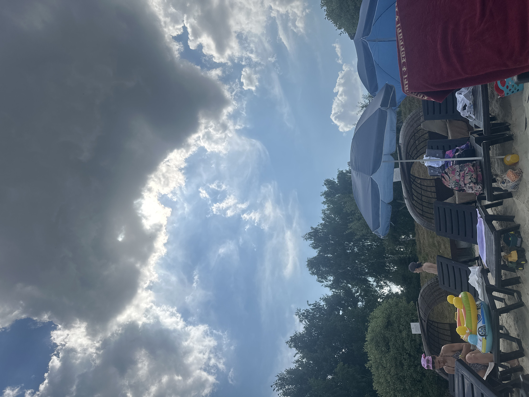 The image depicts a group of people relaxing on a sandy beach under a cloudy sky. The sun is visible, peeking through the dark clouds, suggesting it's daytime. Two blue beach umbrellas are open, providing shade. Several lounge chairs are occupied by people. One person is sitting in a chair, wearing a bikini and a pink headscarf, with a colorful inflatable toy in front of them. Another person, a young boy wearing a baseball cap, is standing near some chairs, facing away from the camera. Beach toys, including a yellow bucket and a toy truck, are scattered on the sand. The scene is set with lush green trees in the background. The weather appears to be partly cloudy. There is a red towel draped over a chair with the word "Ferrari" visible in white text. The overall mood seems relaxed and leisurely, indicative of a summer day at the beach.