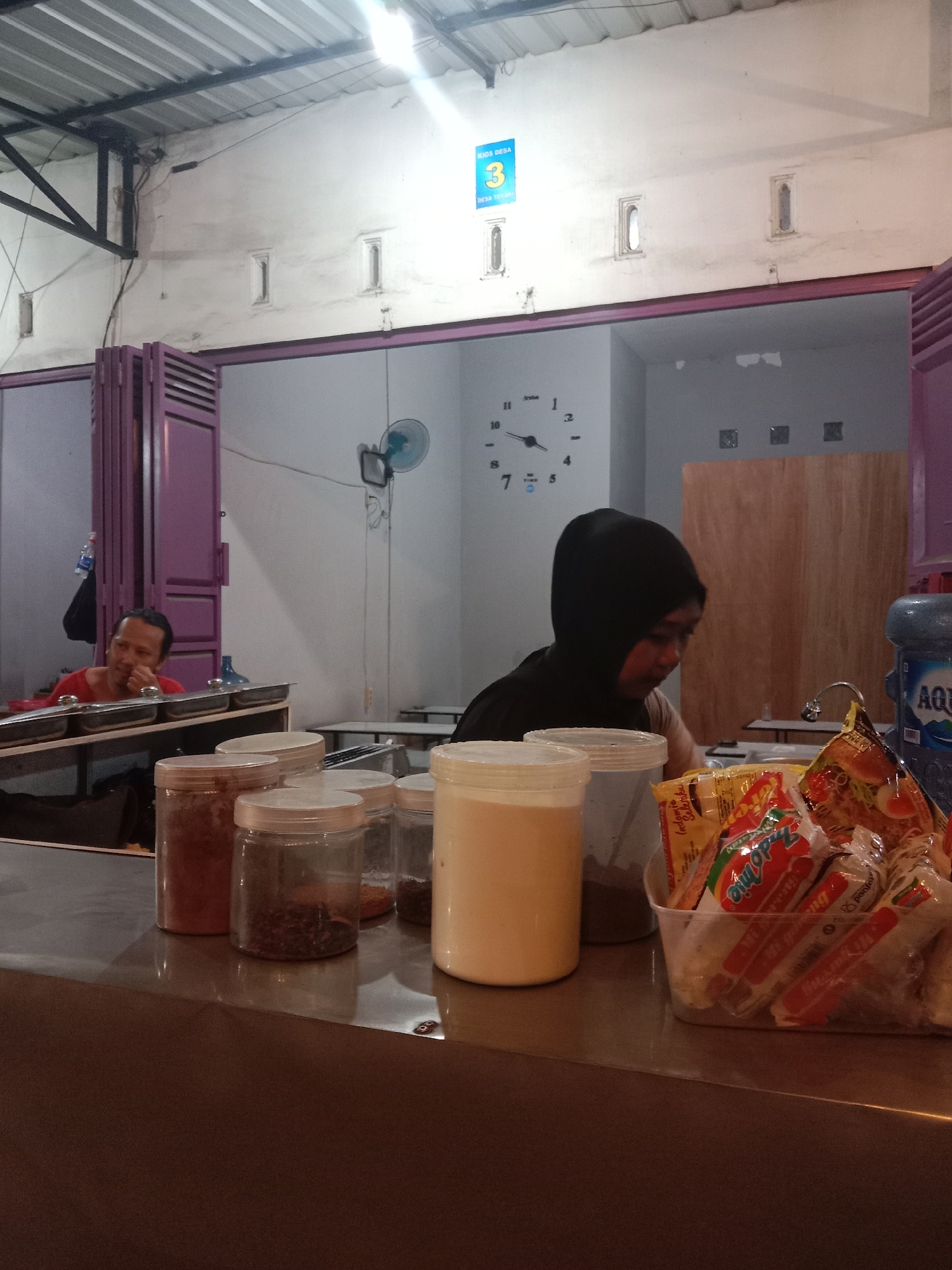 This image depicts the interior of a local eatery or kiosk, identified by a blue sign on the wall as "KIOS DESA 3 DESA TOSINA," within the general location of Cangkring, Indonesia.
In the foreground, a gray counter surface holds several clear plastic jars with white lids, containing various dry goods such as red spices, dark peppercorns, and other granular ingredients. To the right, a large clear plastic container is filled with multiple packets of instant noodles, predominantly showing the "Indomie" brand. A tall, opaque white container stands to the far left.
Behind the counter, a person wearing a black head covering (hijab) is visible from the chest up, facing right and appearing engaged in an activity on the counter surface, though their hands are obscured. To the left and further back, another man in a red shirt is seated, looking towards the left side of the frame, with his right hand near his mouth. He is positioned behind a metal food serving station featuring multiple lidded rectangular trays. A bottle of Aqua mineral water hangs in a black bag to his left.
The environment features light-colored walls, possibly white or light grey, with three small, rectangular, vertically oriented vents or windows visible in the upper section. A large, dark-faced wall clock with Roman numerals (displaying approximately 9:45) is mounted on the wall to the left of the person in the black head covering. A light blue ceiling fan is attached to the wall further right, near a wall-mounted light fixture. Above, a metal roof structure with corrugated sheeting is visible. A large blue Aqua water dispenser bottle stands on the counter to the right of the person in black. The background includes purple bifold doors on the left, an area with a faucet and sink, and simple metal tables and chairs. A wooden panel serves as a partition further in the background. The lighting is bright, likely from overhead sources, illuminating the scene and creating reflections on the counter surface.
