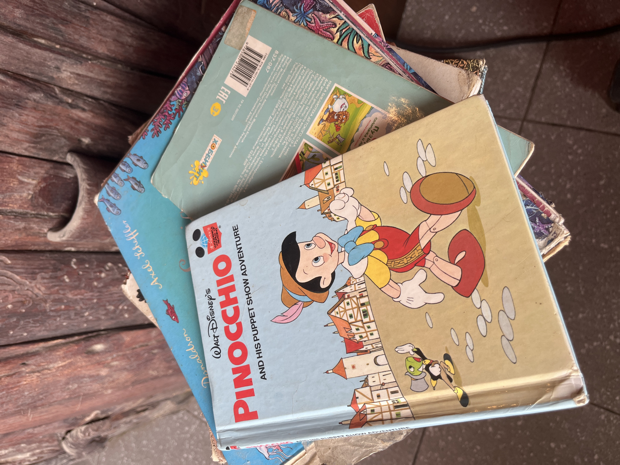 This is a top-down medium shot of a stack of children's books. The primary focus is on a book with a light blue cover titled "PINOCCHIO" in large red letters, with "Walt Disney's" above it in smaller script. Below the title, it reads "AND HIS PUPPET SHOW ADVENTURE." The illustration on the cover depicts Pinocchio in his iconic outfit, with Jiminy Cricket to his left, standing in front of a European-style village. The books are placed on a rustic wooden surface, which appears to be part of a chair or bench. The scene is dimly lit, suggesting an indoor or shaded outdoor environment. The location is specified as ตำบลเกาะพะงัน, Thailand, however, there are no visual elements in the image that specifically indicate this location.