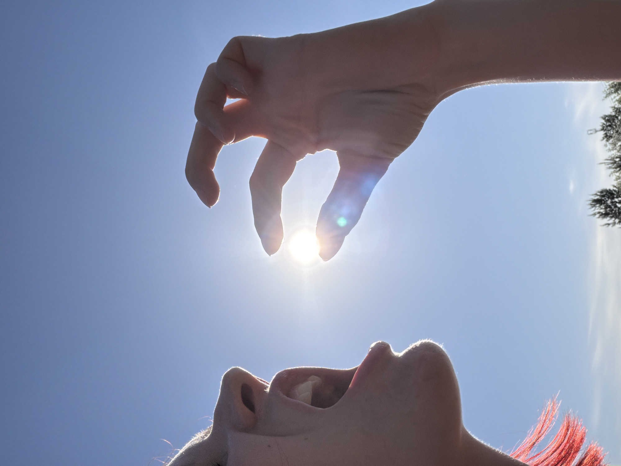 A low-angle, outdoor shot shows a person's head tilted back, mouth open, seemingly positioned to receive the sun held between the fingers of a hand above. The hand is stretched out, the fingers slightly curved. The sun shines between two fingers, creating a bright glare. The sky is clear and blue. Trees are partially visible in the background, slightly out of focus. The subject has red hair and fair skin. The location is unidentified, and it is not possible to confirm the specific location of Chaikovsky, Russia.