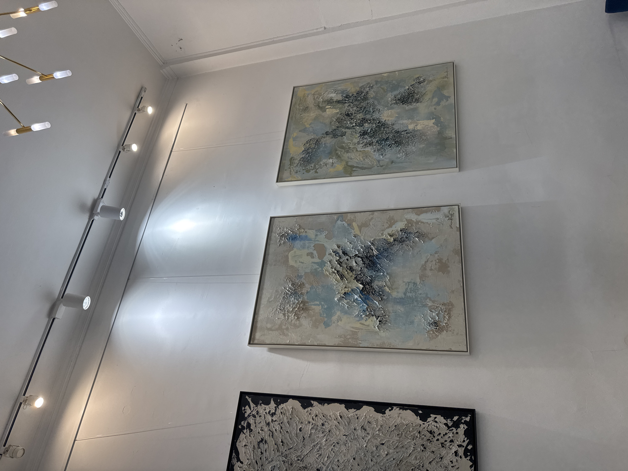 The image shows a wall in an art gallery with three abstract paintings hanging on it. The paintings have textured surfaces and a palette of neutral tones with accents of blue and yellow. The wall is white and textured, with visible imperfections. Above the paintings, a track lighting system is installed, illuminating the artworks. A modern chandelier with multiple lights is also visible in the upper right corner of the image. The overall scene suggests an indoor exhibition space, likely in a contemporary art gallery. This scene is located in the city of Sofia, Bulgaria.